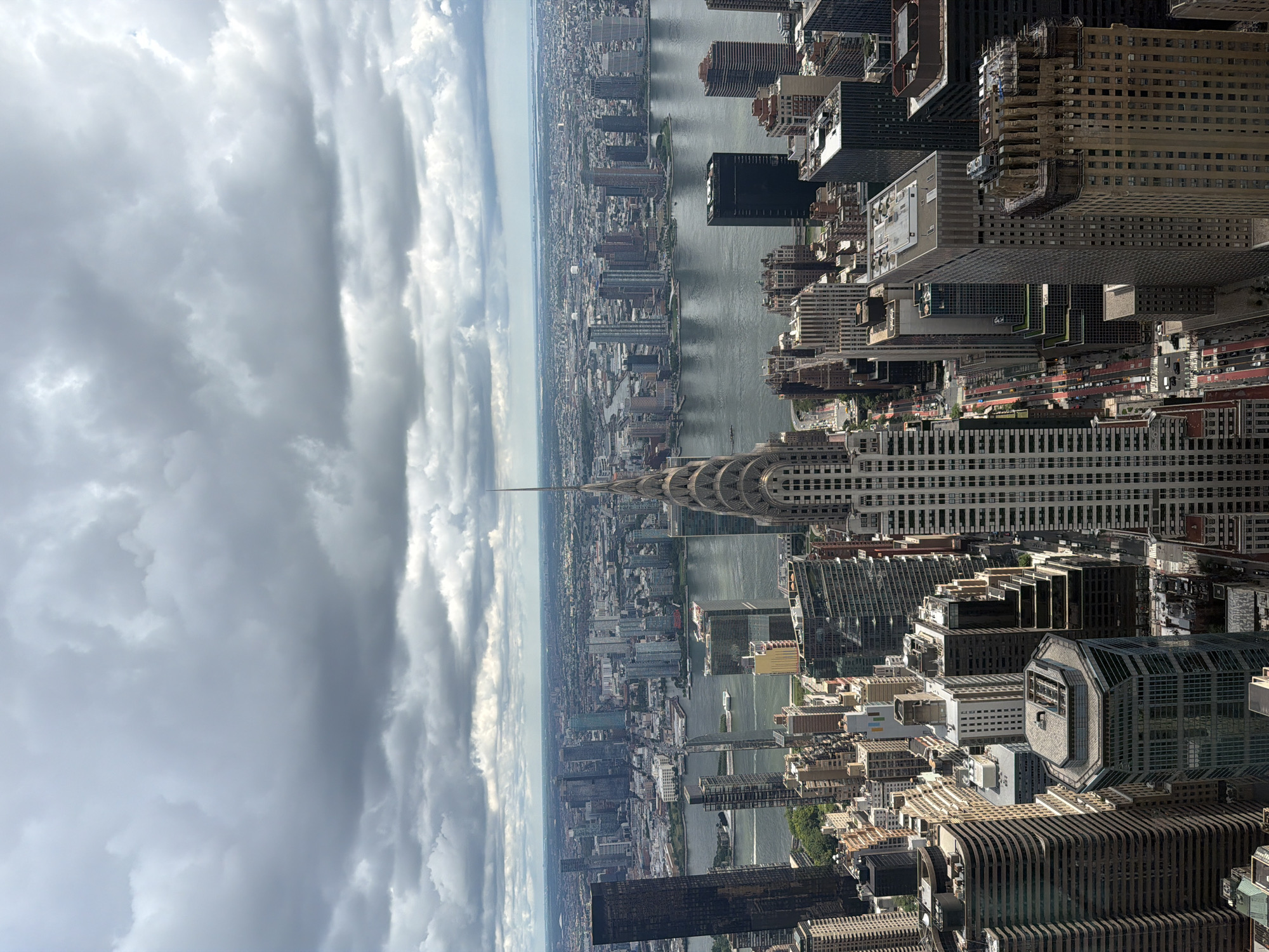 This is a high-angle, vertical, wide shot of the New York City skyline, viewed from above. The Chrysler Building, an iconic Art Deco skyscraper, is prominently featured in the foreground, with its distinctive spire reaching towards the sky. The East River flows between Manhattan and Queens, with buildings and a distant horizon visible on the far side. The sky is overcast with dramatic, dark clouds, suggesting a stormy or moody atmosphere. Numerous other skyscrapers of varying heights and architectural styles populate the scene, creating a dense urban landscape. Some of the buildings are modern, while others appear older, reflecting the city's long architectural history. Traffic is visible on some of the streets below, appearing as red lines on the road. There are no people or animals clearly discernible in the image. The overall impression is one of an immense and complex urban environment under a dramatic sky.