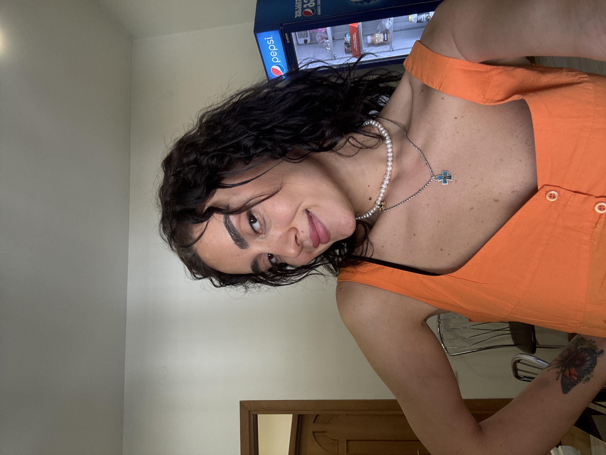 In a casual selfie taken indoors, a young woman with curly dark hair and light skin poses with her left arm extended and hand on her hip. She wears an orange tank top with white buttons down the front and a pearl necklace with a cross pendant. A tattoo of a butterfly is visible on her left forearm. Behind her, to the right, a blue Pepsi vending machine is partially visible, with its door open to reveal various beverages. To the left, a wooden door with a paneled design is ajar, revealing a glimpse of a room beyond. The background is a plain, light-colored wall. The lighting suggests an indoor setting, possibly a commercial space or a room in a building. The image does not contain specific landmarks or features that definitively place it within the city of Tbilisi, Georgia.