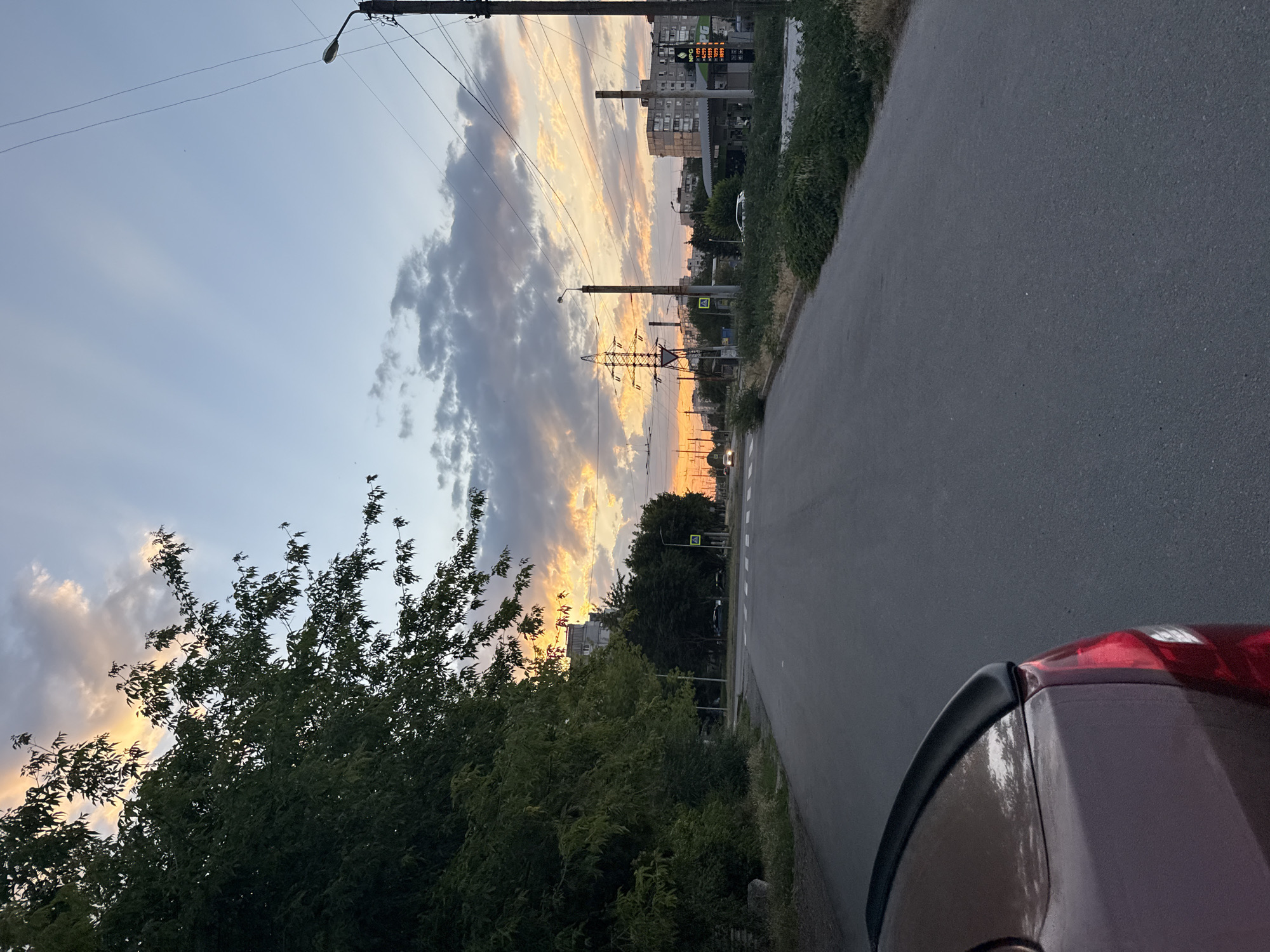 This media file is a landscape photograph taken at dusk. The primary subjects are a street and a dramatic sky filled with clouds illuminated by the setting sun. A portion of a dark-colored car is visible in the foreground, positioned on the right side of the frame.
The scene depicts an urban or suburban environment. In the background, there are buildings, power lines, and streetlights, suggesting a developed area. Trees and dense foliage line the left side of the asphalt road, creating a contrast between nature and infrastructure. The road itself is paved and appears to be a thoroughfare, with a pedestrian crossing visible further down.
The time of day is clearly indicated by the sunset colors painting the sky – vibrant oranges, yellows, and purples streaking through the clouds. The weather appears to be partly cloudy, with the sunlight breaking through to create a beautiful atmospheric effect.
While no people are explicitly visible, the presence of the car and the road implies human activity. The overall mood of the image is serene and contemplative, largely due to the striking sunset and the quiet atmosphere. No visible text is discernible in the image.