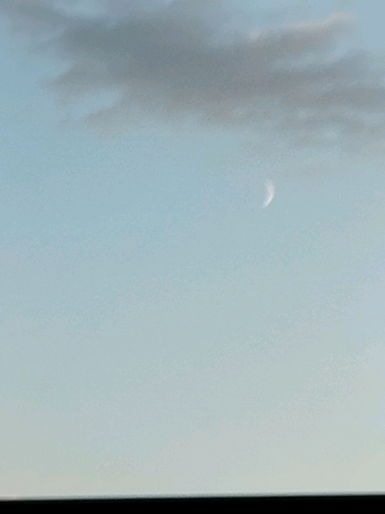 The image shows a pale blue sky with a wispy grey cloud formation in the upper left quadrant. A thin, crescent-shaped white object, possibly the moon or a streak of light, is visible in the upper right quadrant of the sky. The bottom of the image is a solid black band, suggesting the presence of a surface or a border. No people, buildings, or identifiable landmarks of Schiau, Romania are present in the visible portion of the image.