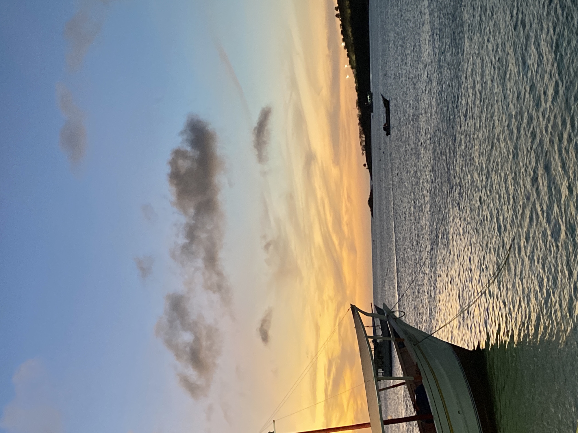 The image presents a tranquil coastal scene at either sunrise or sunset in Juan Griego, Venezuela. The sky, occupying the upper portion of the frame, displays a striking gradient of colors, transitioning from warm yellows and oranges near the horizon to soft blues in the upper atmosphere. Scattered, dark-hued clouds drift across this colorful backdrop.
The lower half of the image features a calm body of water, likely a bay or the ocean. Its surface is subtly rippled, catching and reflecting the ambient light, creating a textured, shimmering appearance. In the foreground, on the right side of the image, the bow and part of the rigging of a boat are visible, silhouetted against the illuminated water and sky. Another smaller, darker boat is discernible further out in the water, positioned towards the distant land.
A dark, narrow strip of land or an island forms the horizon line, silhouetted against the vibrant sky. Several small, bright lights punctuate this distant landmass, suggesting human settlement or infrastructure. The overall atmosphere of the scene is one of serenity and peace. There is no visible text in the image.