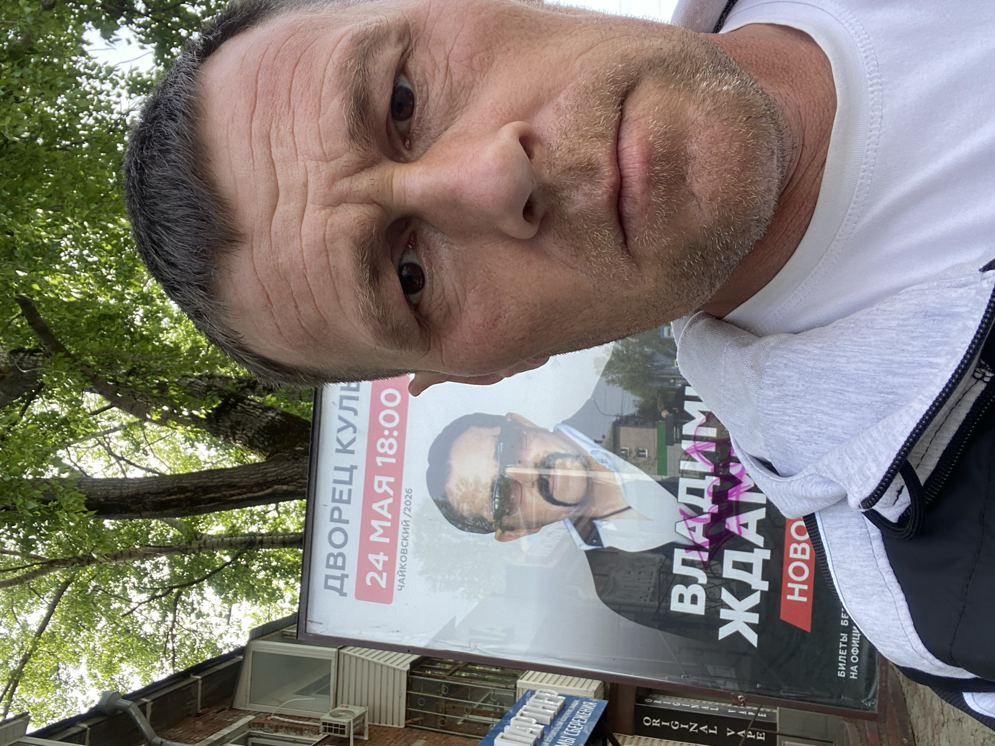 A selfie is captured from a high angle of a Caucasian man with close-cropped graying hair, wrinkles on his forehead, and stubble on his face. He is wearing a white t-shirt and a gray and black hooded jacket. Behind him is a campaign poster advertising a performance by Vladimir Zhdanov on May 24th at 18:00 at the Palace of Culture in Chaikovsky. The poster includes the year "/2026", which could suggest a promotional connection with the city's anniversary. The poster is in Russian, and the bottom corner of the poster lists "ORIGINAL VAPE." Above the poster, tree branches with green foliage are visible, suggesting an outdoor location. The environment includes a building with an air conditioning unit.