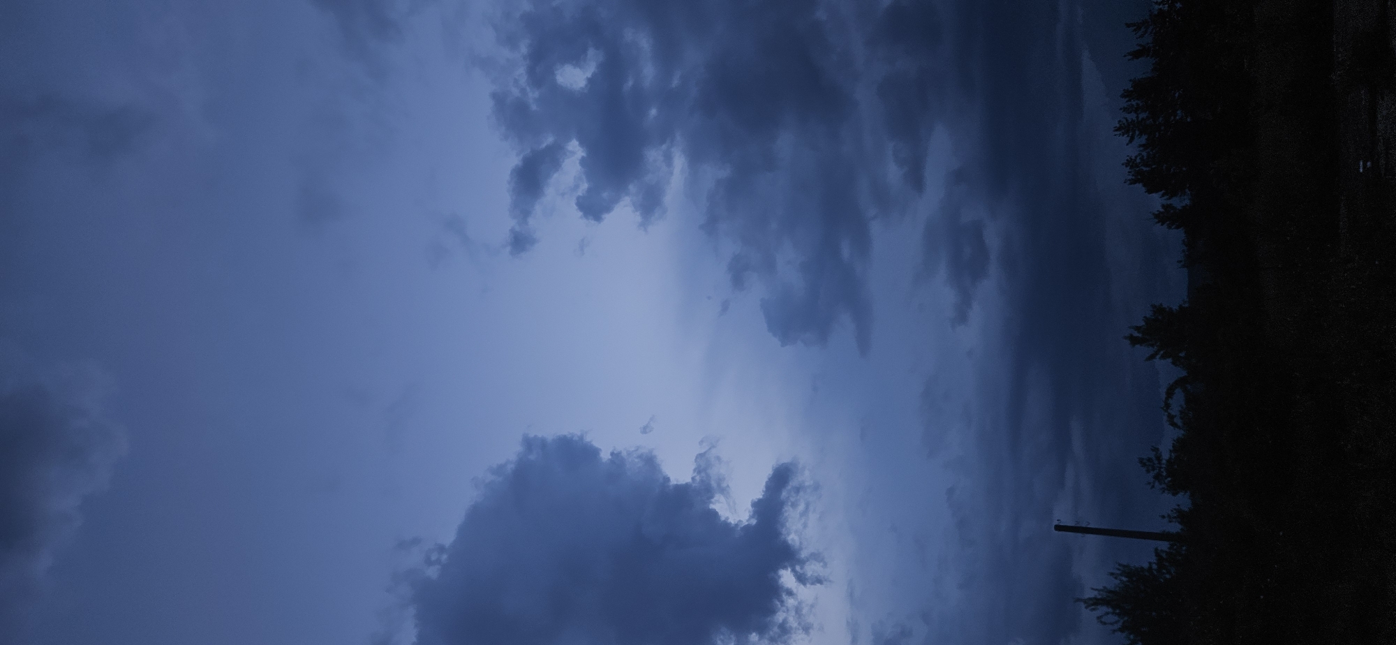 The image captures a dramatic, dark sky dominated by heavy, deep blue and grey clouds, suggesting pre-storm conditions or twilight. The sky is largely overcast, with dense cloud formations stretching across the frame. Lighter patches of blue are visible through and between the clouds, particularly towards the upper-left and central areas, hinting at a distant or obscured light source.
Along the right side of the image, a dense silhouette of trees and foliage forms a dark, irregular horizon line. Amidst these dark outlines, a single utility pole stands vertically, also appearing as a dark silhouette against the dimly lit sky. The lighting suggests either very late evening, early morning, or a heavily overcast day with an impending storm. The overall mood is somber and quiet, characteristic of such weather conditions. No specific human or animal activity is visible. There is no visible text in the image. The setting is Gutsal, Bulgaria.