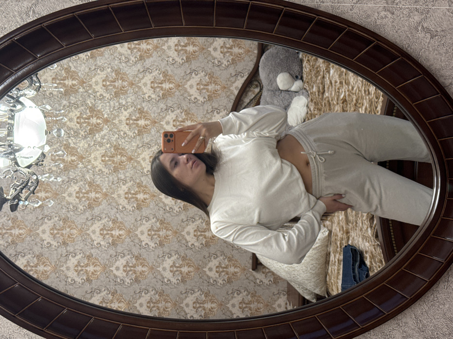 A female subject is depicted taking a mirror selfie indoors in a room located in Vysokinichi, Russia. The subject, possessing long, dark hair, is wearing a white long-sleeved cropped top and light grey drawstring sweatpants, exposing her midriff. She holds a smartphone with an orange case in her right hand, directed towards the mirror, with a ring visible on her right ring finger. Her left hand rests on her hip. The image reflects an oval-shaped mirror with a dark brown, segmented frame. The background visible through the mirror shows a wall covered with ornate patterned wallpaper featuring light brown and grey tones. A chandelier with multiple lights and crystal-like elements hangs from the ceiling. A bed with a dark wooden headboard and a light brown textured bedspread is partially visible in the lower portion of the reflection. A patterned pillow and a pair of folded blue jeans are present on the bed to the left, while a large grey stuffed animal lies on the bed to the right. A dark wooden nightstand is partially visible to the left of the bed.