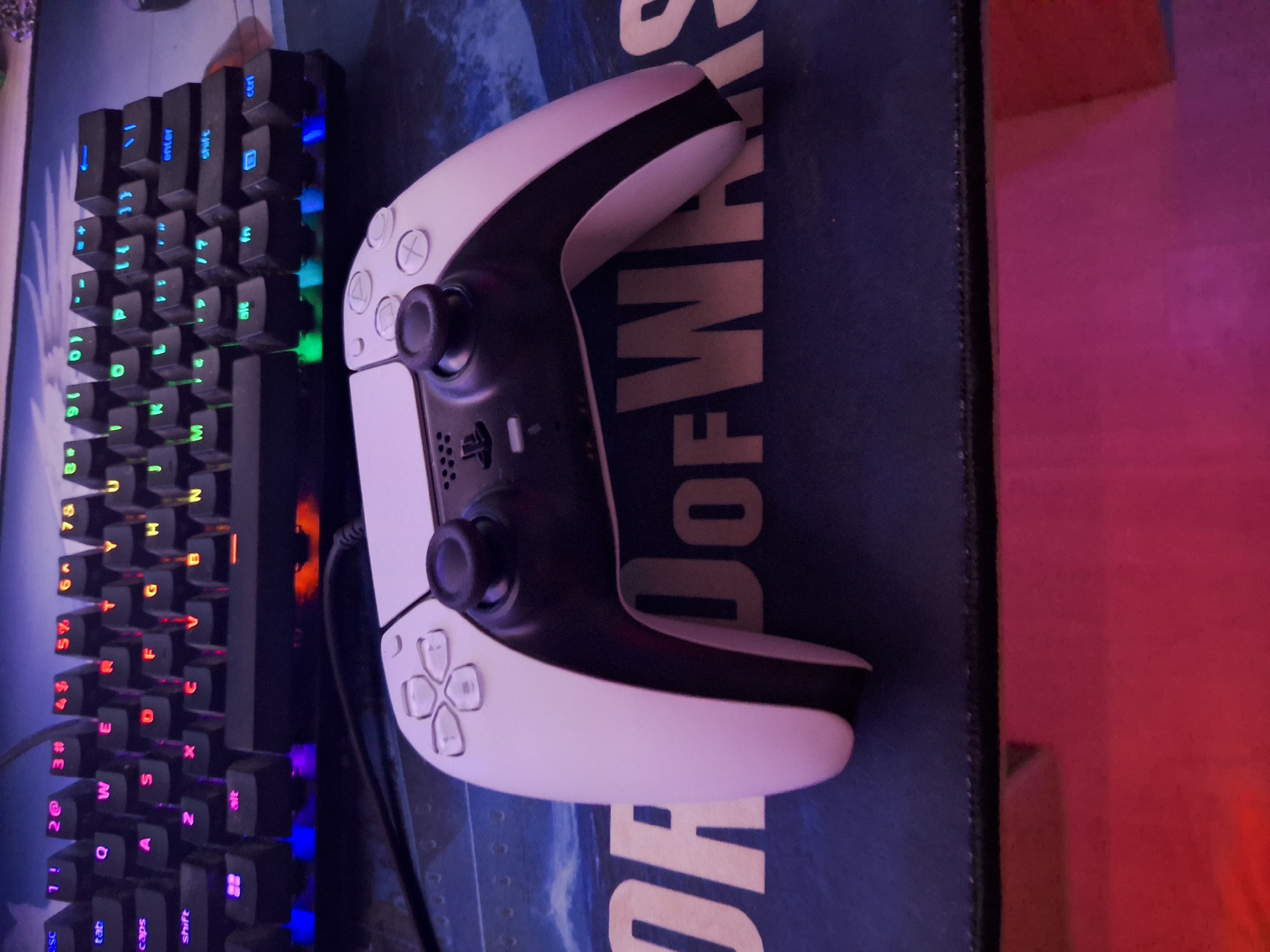 An overhead shot captures a white and black video game controller placed on a dark blue mousepad. The mousepad features the words "OF WARS" in large, light brown lettering, partially obscured by the controller. Behind the mousepad, a black keyboard with illuminated keys is visible. The keys display a variety of colors, including pink, orange, blue, and green. The background is dark and out of focus. The scene is lit with ambient light, creating a reddish glow on the bottom edge of the image. No people or animals are present in the scene. The image appears to be taken indoors, possibly in a gaming setup.