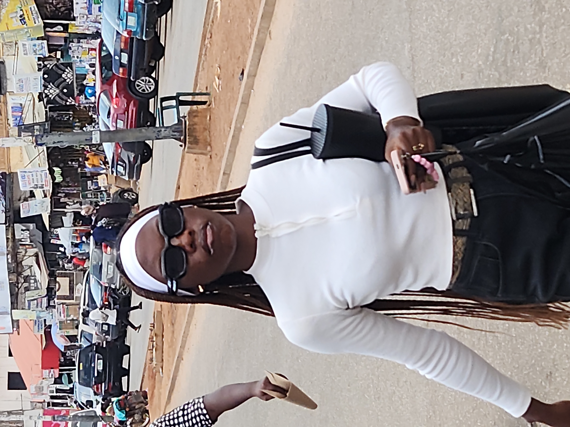 A full-shot, eye-level outdoor street scene shows a young woman walking towards the camera. She is dressed in a white, long-sleeved, collared top with a zipper at the neck, and dark denim jeans with a patterned belt. She wears black sunglasses and a white headband. In her right hand, she holds a pink smartphone and in her left hand, a black tumbler cup with a straw. She also carries a black shoulder bag. Behind her, the street is lined with various market stalls and shops, with a variety of vehicles, including cars and a motorcycle, parked or driving on the road. The background appears to be a bustling urban environment.