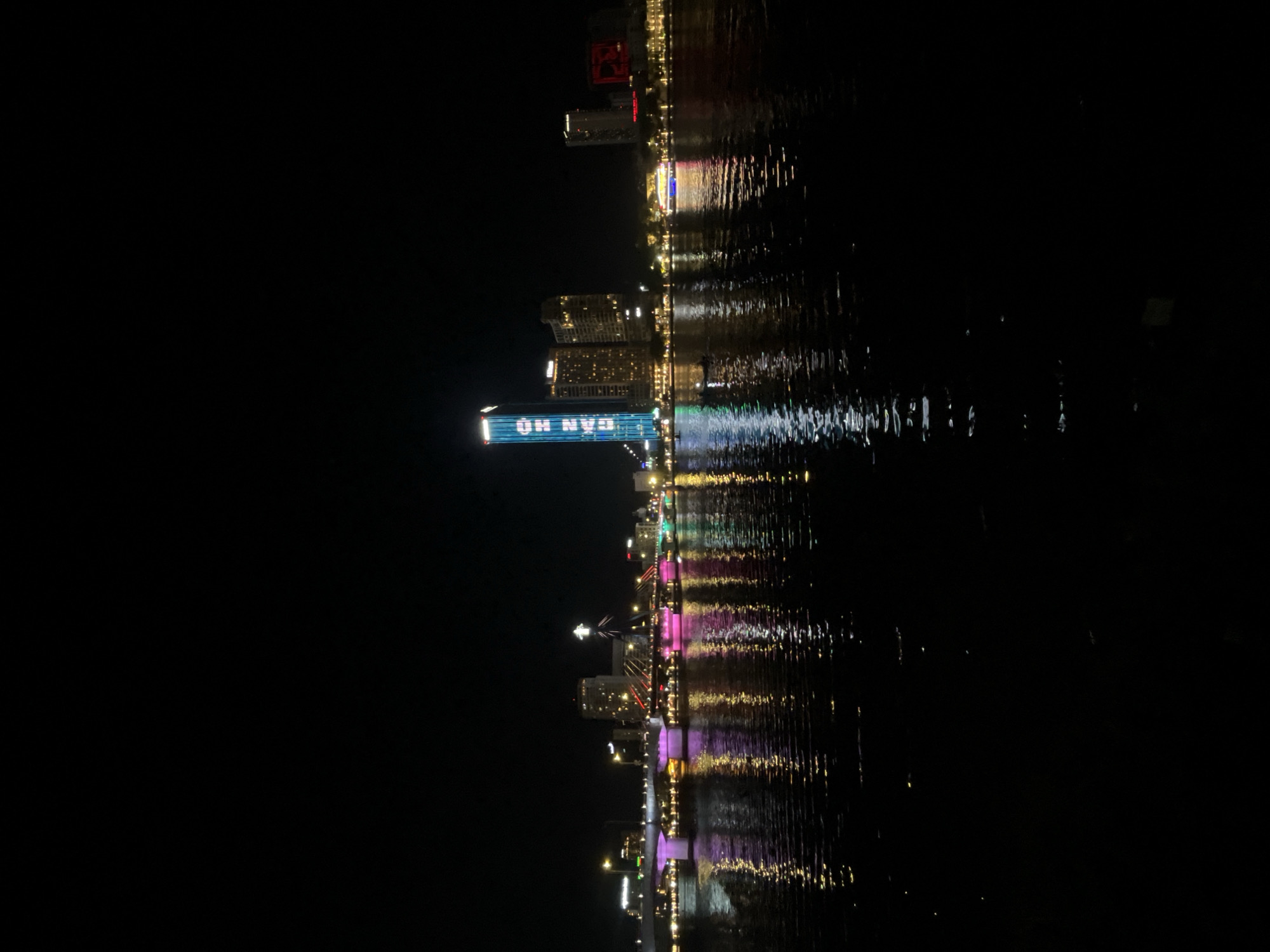 This is a night photograph of the Han River in Da Nang, Vietnam. The city skyline is illuminated by various lights, with a prominent skyscraper displaying blue text. Several bridges are visible, adorned with colorful lights reflecting on the water. The river itself is dark, with shimmering reflections of the city lights. There are no people or specific actions discernible in the image.