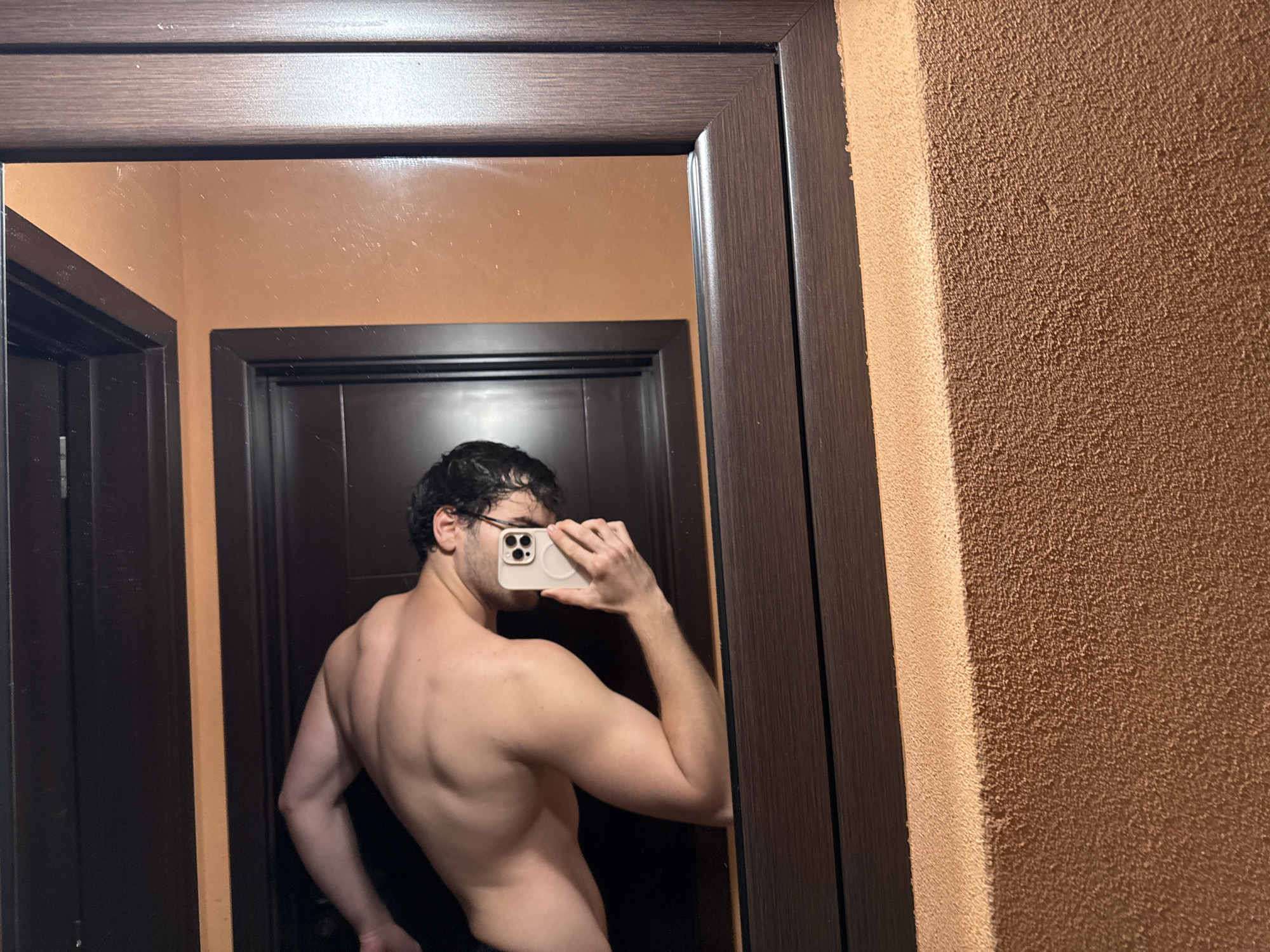 A person with dark hair and glasses is holding a smartphone and taking a photo of themself in a mirror. The person's upper body is bare. The mirror is framed with a dark brown wood finish and reflects a dark doorway and an orange-colored wall. The adjacent wall to the mirror is a textured, brown color. The location cannot be determined from the image.