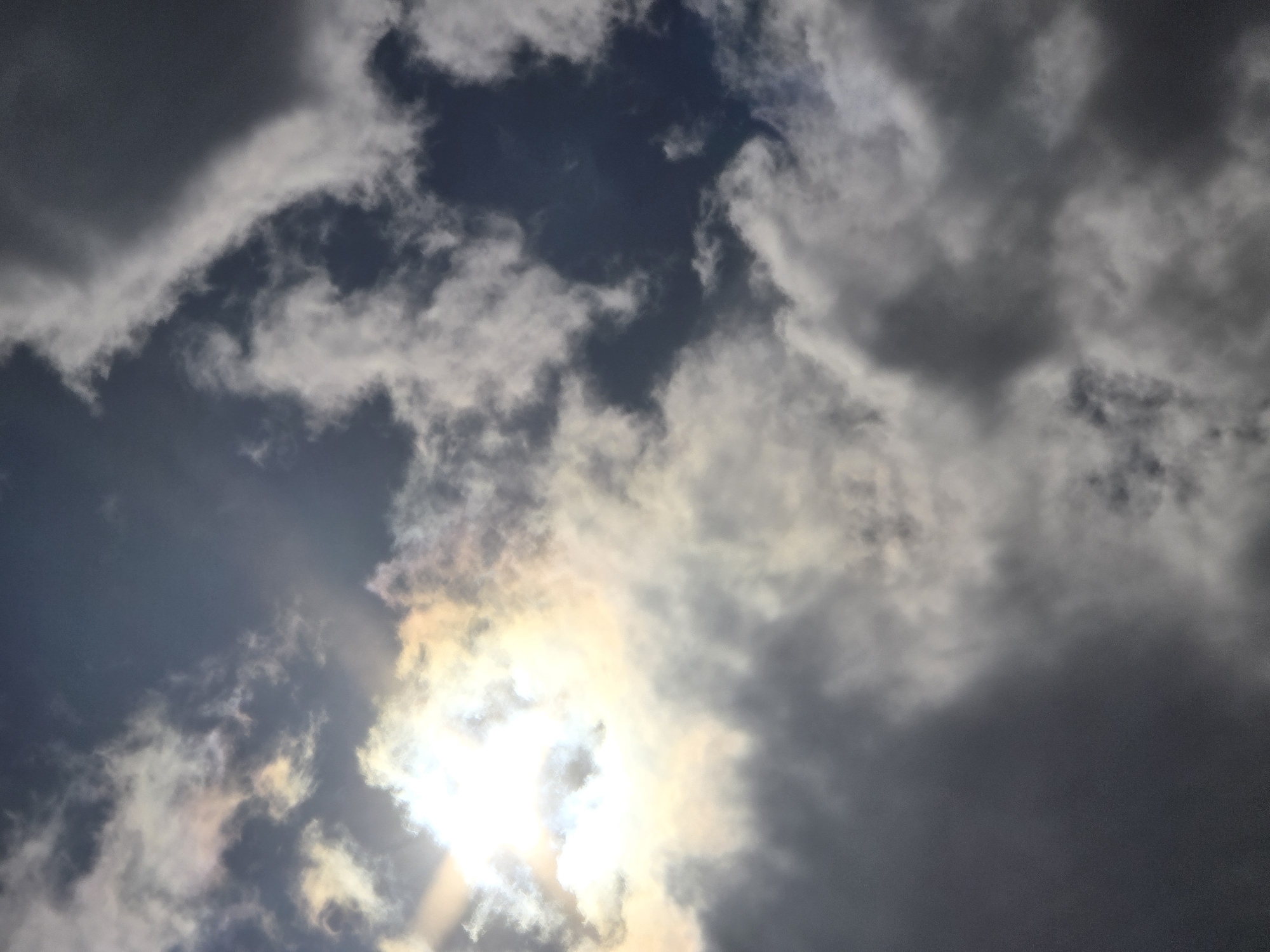 This image depicts a sky filled with dramatic clouds, with the sun partially obscured by them, creating a luminous effect. The clouds range from bright white to dark gray, suggesting varying atmospheric conditions, possibly indicating an approaching storm or recent rainfall. The sun's rays are visible, casting a soft glow and creating subtle colorations within the clouds, hinting at potential iridescence. The scene is a purely natural one, focused on the sky. No specific urban features of Sofiivska Borshchahivka, Ukraine, are visible in this close-up view of the sky. There are no people, objects, or actions to report.