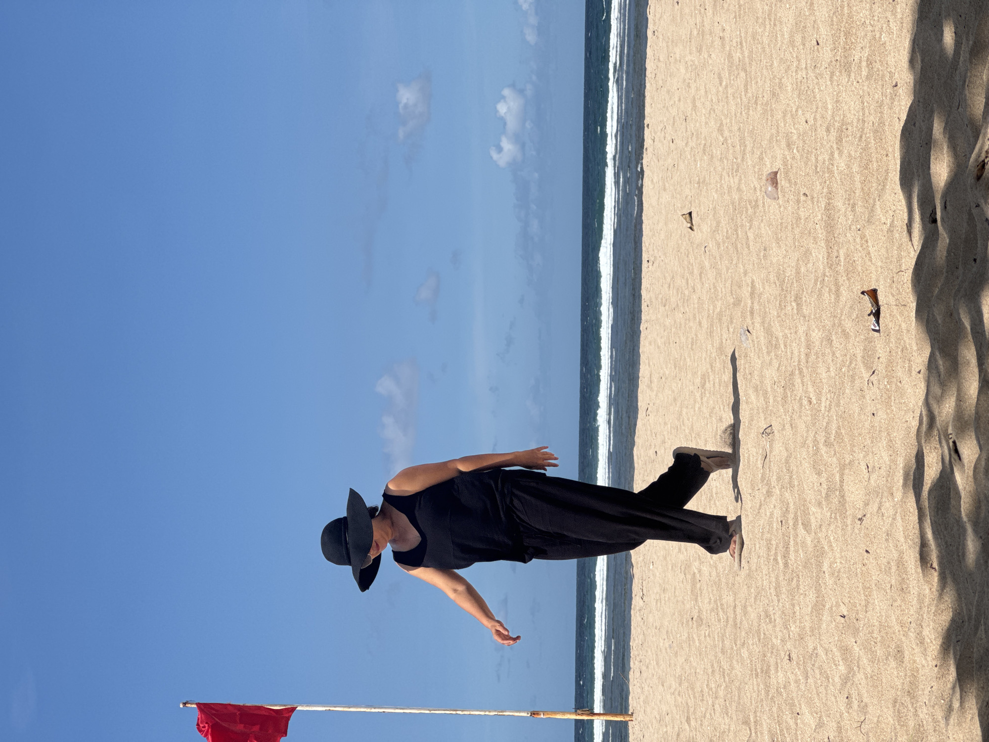 A person wearing a black hat and black clothes walks on the beach in Kuta Selatan, Indonesia. The beach is sandy and has a red flag on a pole. The ocean is in the background. The sky is blue with some clouds.