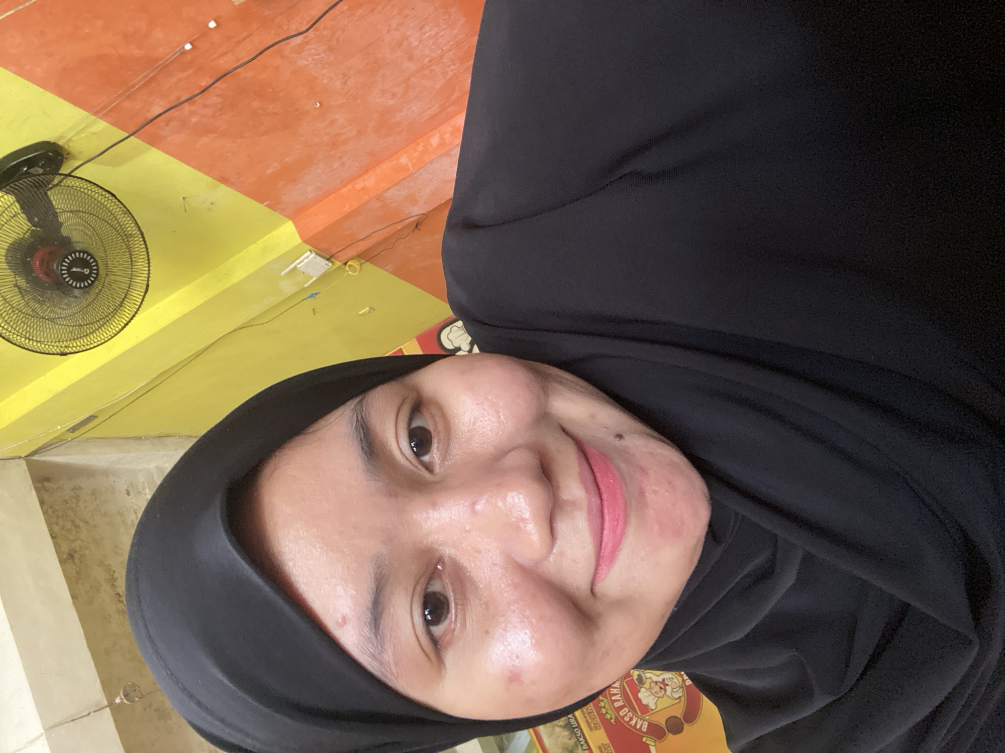 The image shows a close-up, selfie-style shot of a young woman wearing a black hijab. She is smiling and looking at the camera. Her face has some visible blemishes. Behind her, a wall is painted in diagonal sections of yellow and orange. A black oscillating fan is mounted on the upper part of the yellow section. To the left of the woman, part of a circular yellow sign with red lettering and an illustration of a chef is visible. This sign appears to be associated with a food establishment, possibly a restaurant or food stall. The overall environment suggests an indoor setting, potentially a casual dining area in Sungai Sipai, Indonesia.