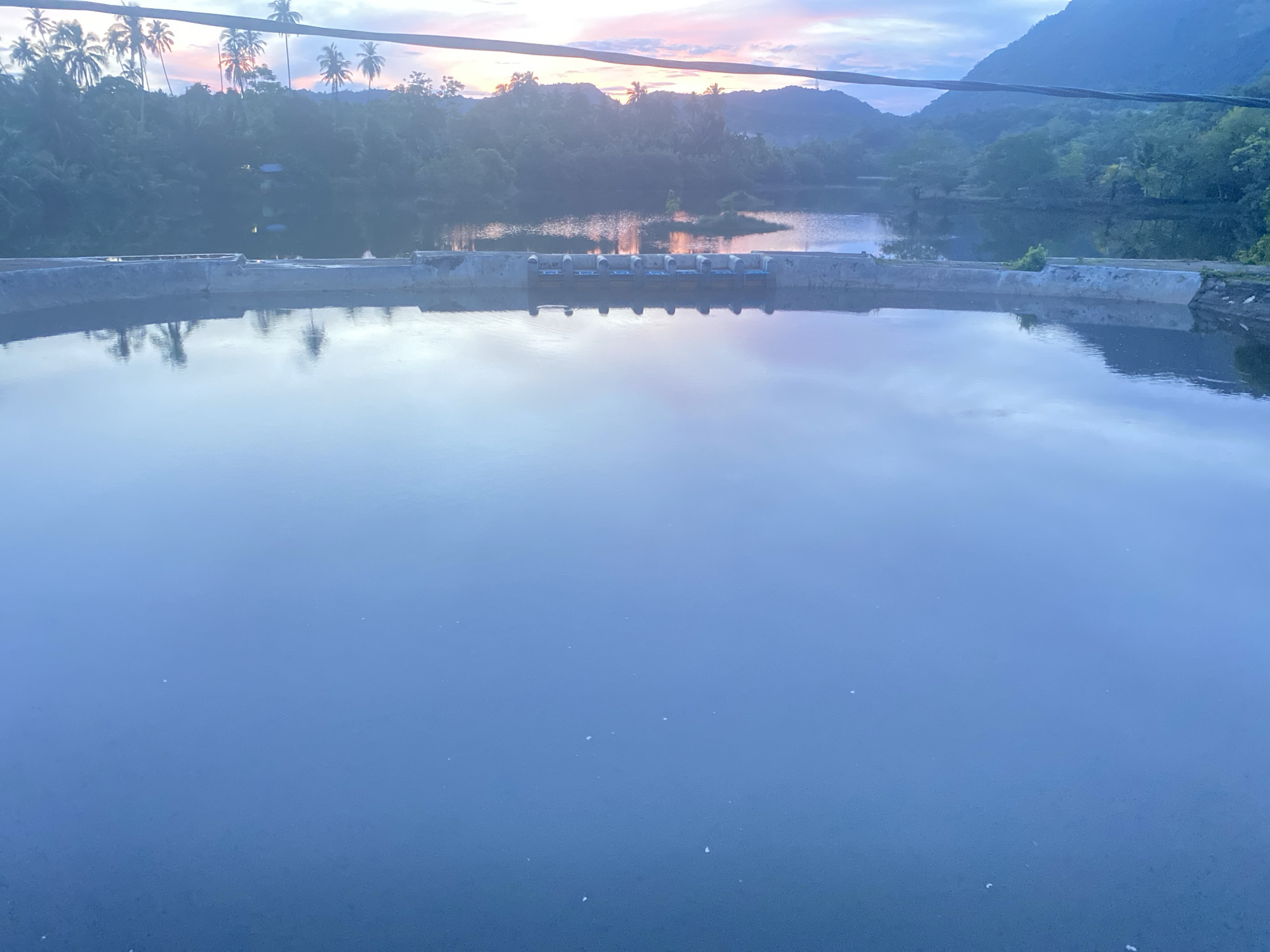 The image shows a calm body of water, likely a reservoir or pond, with a concrete structure running across it. The structure has several openings or gates. In the background, there are lush green trees and hills, with palm trees visible on the left. The sky is a gradient of blue and pink, suggesting either sunrise or sunset. There are no people or animals visible in the image. The overall scene is tranquil and natural. The location of Aneuk Paya, Indonesia, is provided contextually but is not directly depicted within the visual elements of the image.