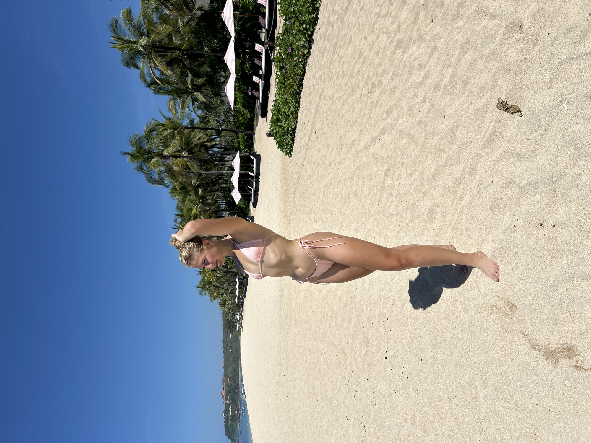 A young woman in a pink bikini stands on a sandy beach. She is fair-skinned and has blonde hair pulled up in a bun. The sky is clear and blue. Palm trees and white beach umbrellas line the beach in the background. The location is likely Kuta Selatan, Indonesia.