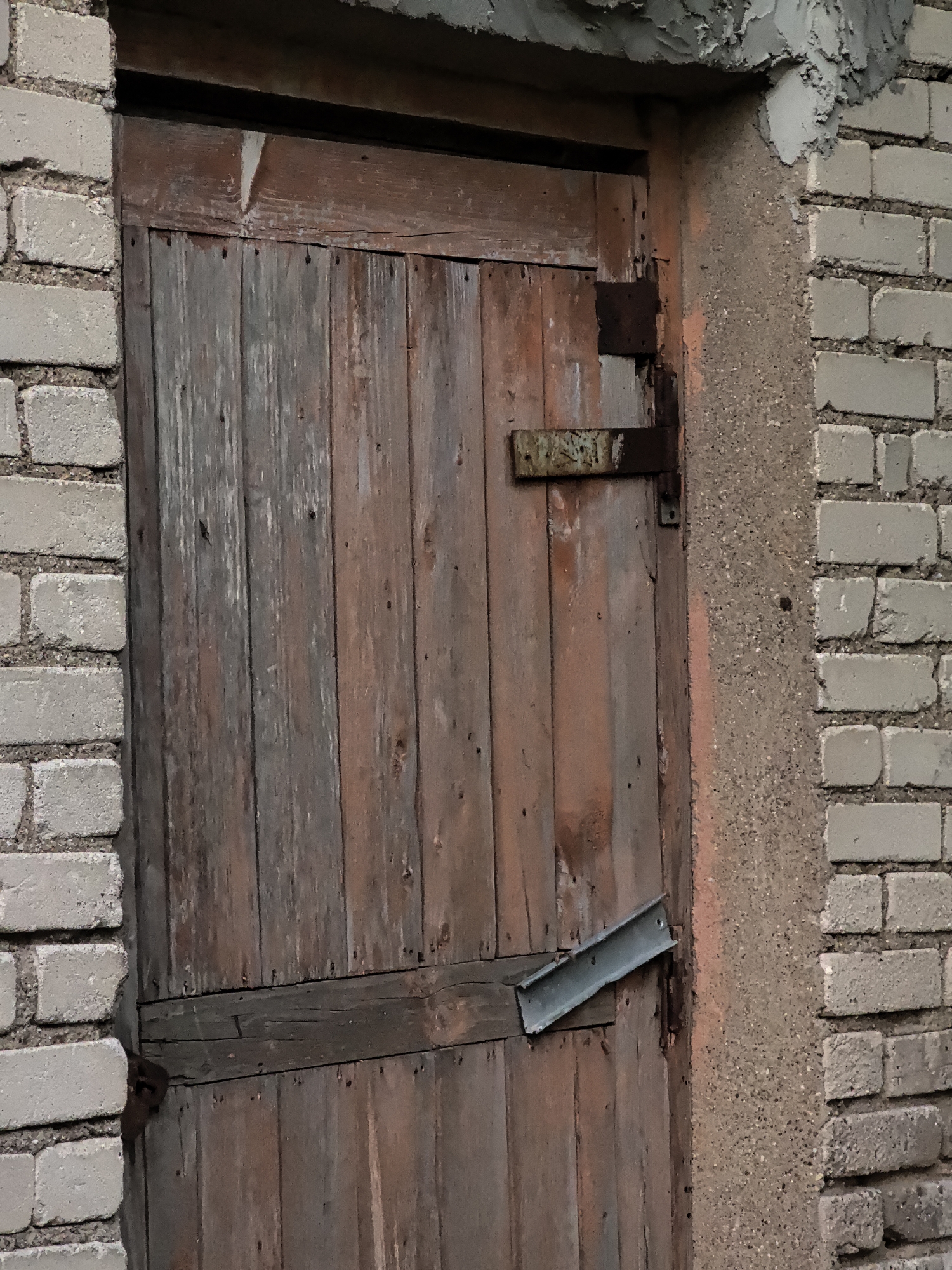 A weathered, wooden door, constructed from vertical planks of varying shades of brown and gray, is set within a rough, textured, peachy-pink frame that abuts a light gray brick wall. The door features two horizontal wooden braces, one near the top and another lower down, with the lower brace showing a metal reinforcement strip attached diagonally across it. A rusty metal hinge is visible on the left side of the door, connecting it to the frame. Above the door, the building's facade is rough, chipped concrete. The scene depicts an exterior view of an old building, likely a shed or utility structure, in an urban environment. No people are present.