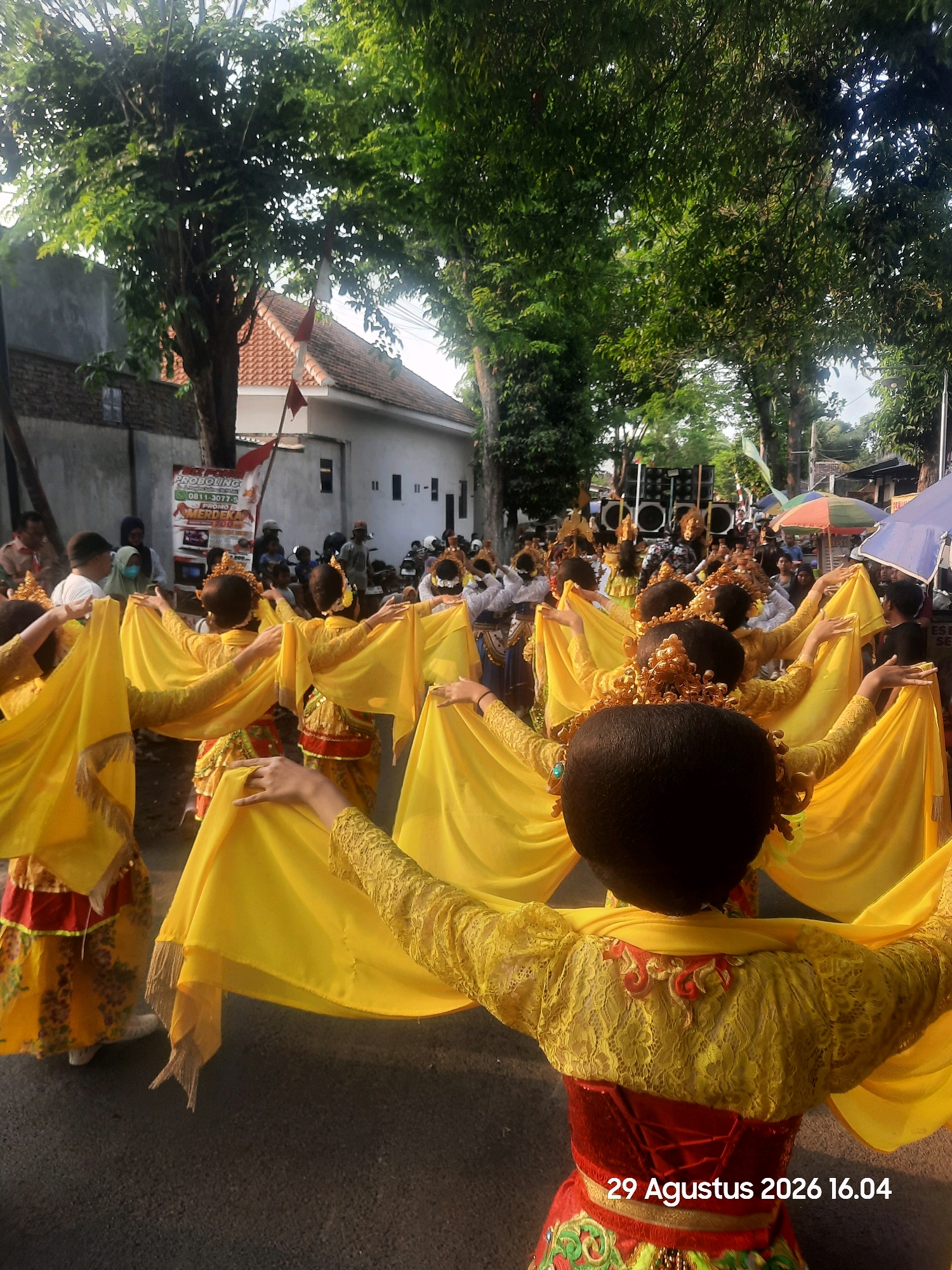 In Patokan, Indonesia, a group of dancers in traditional yellow costumes with intricate gold headpieces are performing outdoors. The dancers are seen from behind, with their arms extended and holding flowing yellow fabric, creating a visually striking effect. They are wearing elaborate yellow lace tops adorned with red detailing and red corsets. The backdrop features trees lining a street, with buildings and a crowd of spectators visible in the distance. There are also speakers on a platform, suggesting a public event or performance. Red and white Indonesian flags are present, adding to the festive atmosphere. The image is dated "29 Agustus 2026 16.04".