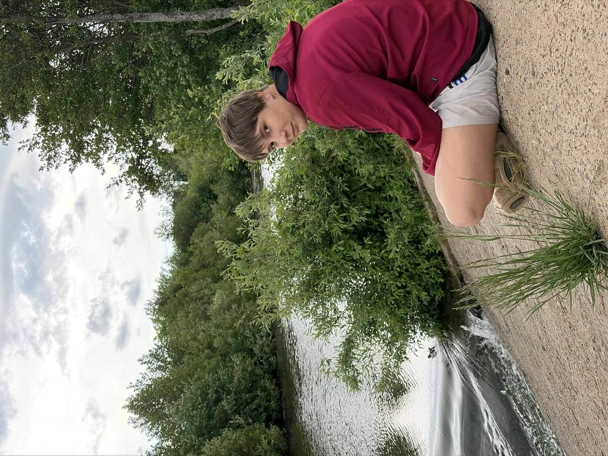 A photograph taken outdoors depicts a young person crouching at the edge of a concrete structure beside a body of water. The young person, facing the camera, is wearing a maroon hooded jacket and light-colored shorts with tan shoes. They are positioned on the right side of the frame. The concrete structure serves as a boundary, likely for the water. To the left, the water is surrounded by dense green vegetation, appearing to be trees and bushes. The sky is visible at the top left, showing overcast conditions with clouds. The image is oriented so that the ground appears on the right and the sky on the left. The location is Nizhnii Vishur, Russia.