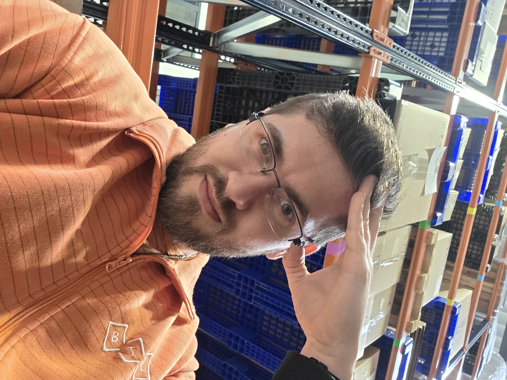 A man with short dark hair, a beard, and glasses is holding his forehead with his hand, looking directly at the camera. He is wearing an orange zippered sweatshirt with white lettering on the lower left. The background shows an indoor warehouse setting with orange and blue shelving units and boxes. The location appears to be a storage facility or similar industrial setting, but there is no specific information suggesting it is in Plovdiv, Bulgaria.