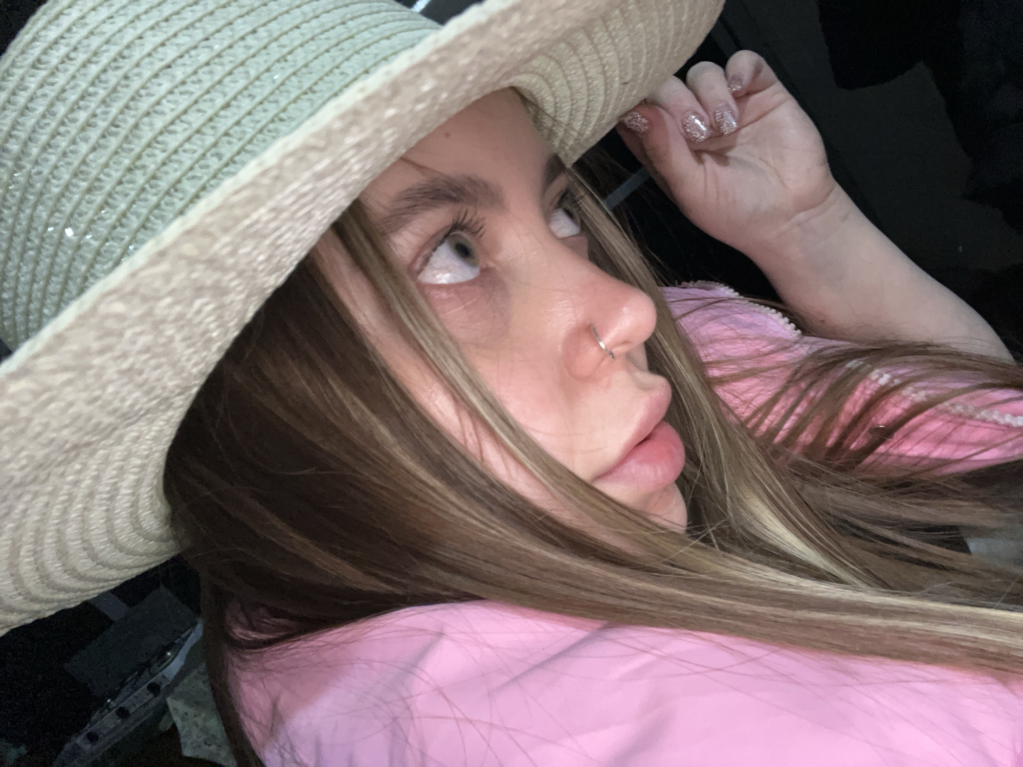 A person wearing a light-colored straw hat and a pink top is looking upwards. They have light skin, brown hair with blonde highlights, and a nose ring. One hand is visible near the hat. The background is dark and indistinct. The city of Novopokrovskaya, Russia is not discernible in the image.