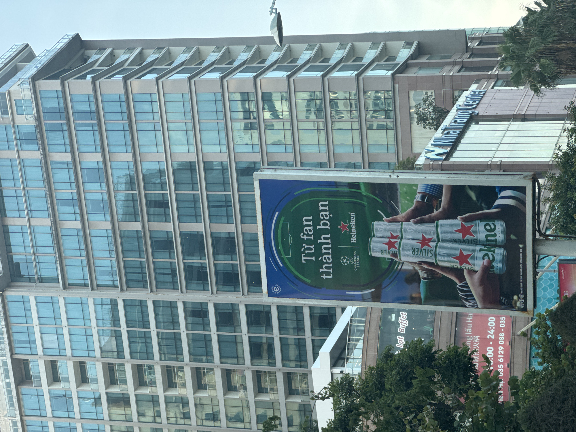 The image captures an urban scene in Nha Trang Ward, Vietnam, on an overcast day. A prominent vertical billboard stands in the foreground, displaying a Heineken Silver advertisement. The billboard features the text "Từ fan thành bạn" (From fan to friend), the UEFA Champions League logo, and a visual of two hands holding four stacked silver Heineken cans. Behind the billboard, a multi-story building with the illuminated "Nha Trang Center" sign in blue letters is visible to the right, with several palm trees in front and to its right. Further left and partially obscured by green foliage, another building displays a large red sign with Vietnamese text, including "Lẩu Buffet" (Hotpot Buffet), "10:00 - 24:00" (operating hours), and a phone number. The background is dominated by a tall, modern high-rise building with numerous floors, characterized by light-colored facades and extensive light-blue tinted glass windows, many reflecting the sky. Some sections of this building feature balconies with glass railings. A modern, curved street light is partially visible in the upper right portion of the frame.