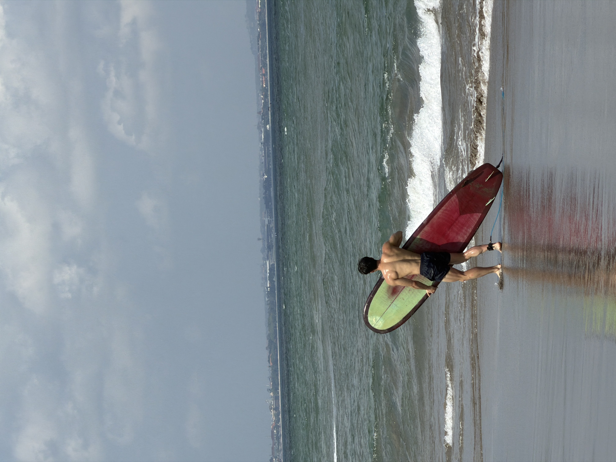 A full-shot vertical, eye-level photograph captures a shirtless, muscular man standing on a wet, sandy beach in North Kuta, Indonesia. He is facing away from the camera, holding a red and green surfboard horizontally with his right hand near the tail and his left hand further up the board. The man is wearing black shorts, and his wet skin glistens in the diffused light under a cloudy sky. He stands at the edge of the water, with gentle waves lapping at his ankles. The ocean, a deep green with white foam cresting, stretches out towards the horizon, where a distant cityscape of buildings and structures is visible under a hazy sky. The wet sand reflects the man and the surfboard.