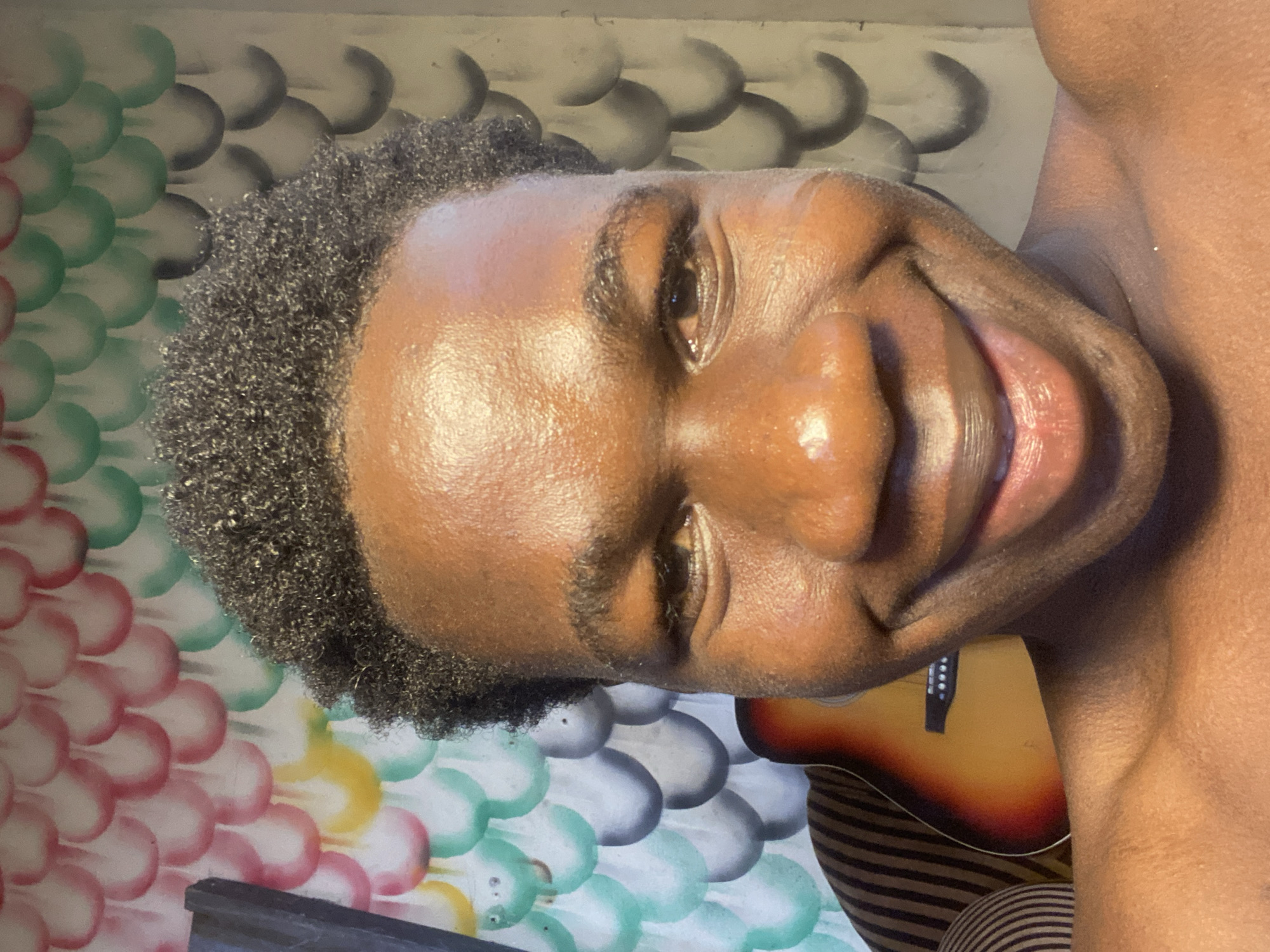 A close-up, portrait-oriented photograph shows a smiling, dark-skinned young man with short, curly black hair. The man's face is well-lit, revealing details of his skin texture and facial features. Behind the man is a wall painted with a pattern of overlapping, scale-like shapes in various colors including white, grey, pink, green, and yellow. A guitar is partially visible in the bottom right corner of the image, resting against a striped cushion. The image provides no definitive clues to confirm a specific location in Mararaba, Nigeria.