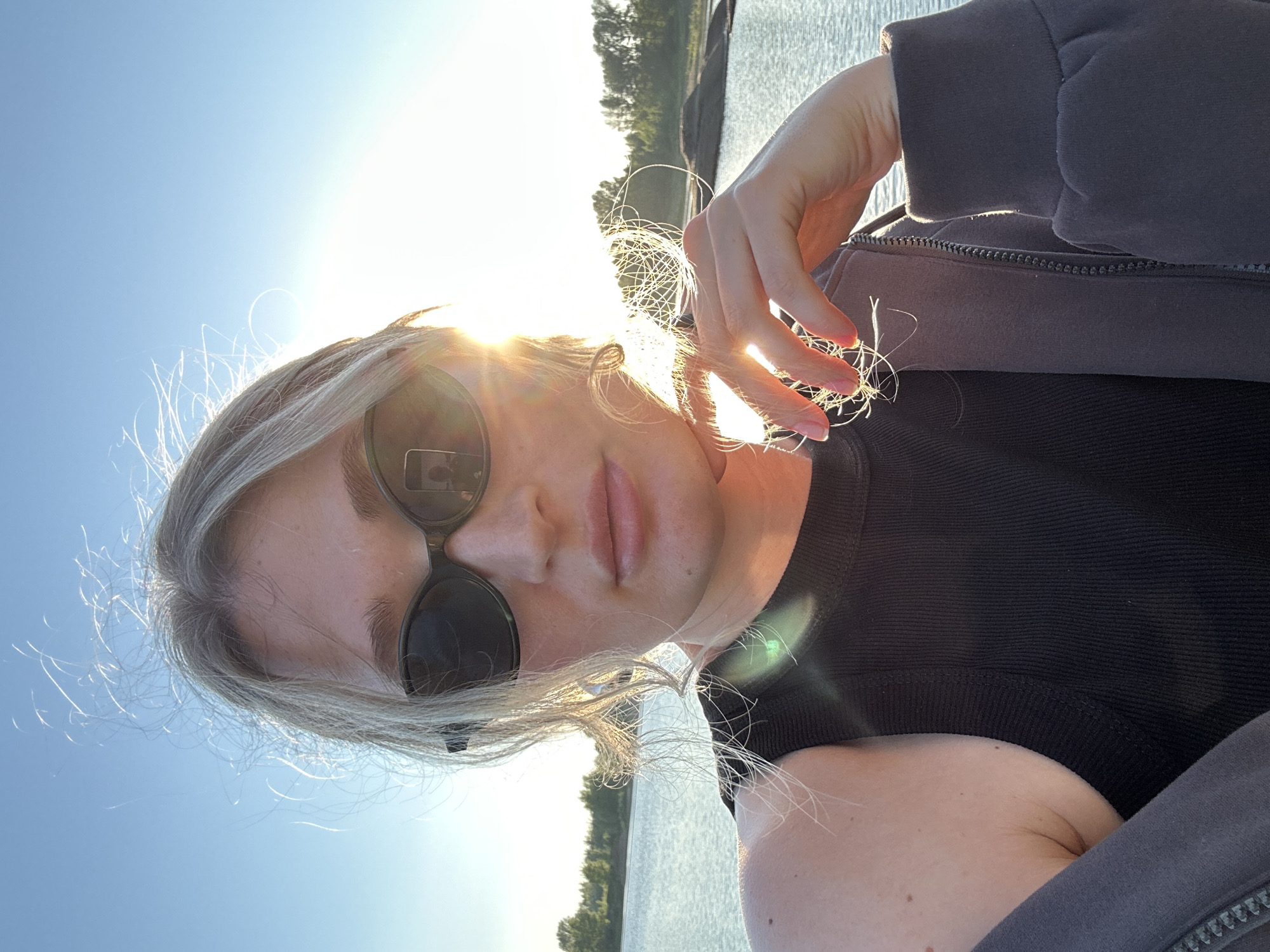 A young woman with blonde hair and sunglasses takes a selfie by a body of water, likely the Kama River, given the location of Chaikovsky, Russia. The sun is setting or rising, casting a warm glow and lens flare across the image. She is wearing a black tank top and a grey hooded jacket. Her hand is near her face, touching her hair. The background shows a calm body of water with a tree line in the distance.