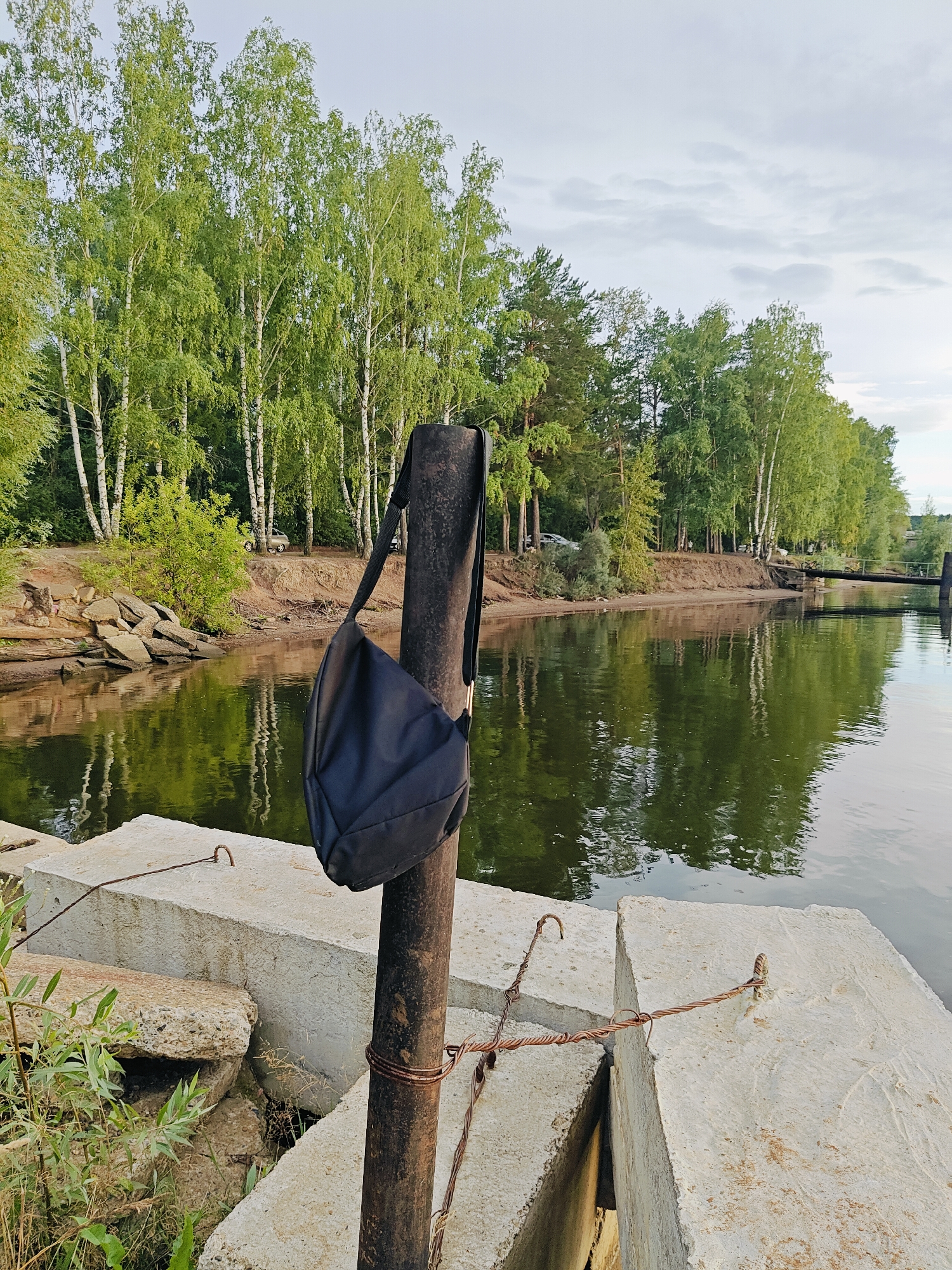 The image depicts a serene riverside scene in Chaikovsky, Russia, likely during daylight hours with overcast skies. In the foreground, a dark-colored shoulder bag hangs from a dark, weathered metal pole. The pole is secured by twisted wire. The bag appears to be made of fabric and is empty. To the left and front of the pole are large, rough-textured concrete blocks, some of which are stained and weathered. One block has a metal hook embedded in it, with rusted wire extending from it. Some dry vegetation and green leaves are visible near the base of the concrete blocks. The background features a tranquil body of water with reflections of trees and the sky. The opposite bank of the river is lined with a dense growth of trees, primarily birch trees with white bark and lush green foliage, interspersed with pine trees. A sandy embankment is visible along the river's edge, with some scattered rocks. A small bridge or pier structure can be seen in the distance across the water. The overall impression is one of a quiet, natural setting.