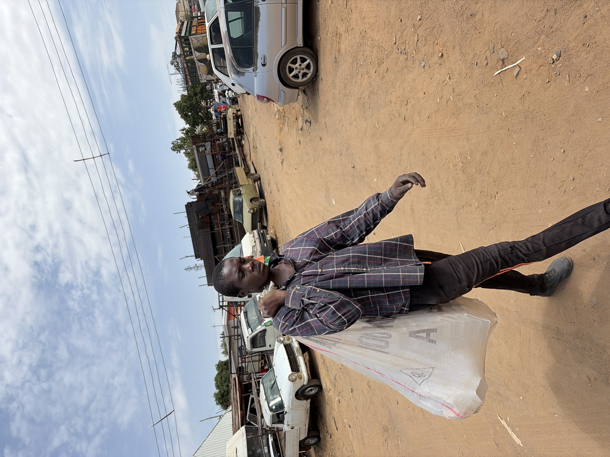 A man stands on a dirt road in Gura Top, Nigeria. He carries a large white bag over his shoulder, and wears a plaid shirt and dark pants. The background shows a street with parked vehicles, including cars, vans, and trucks. Overhead power lines are visible against the sky. Buildings and trees also populate the scene.