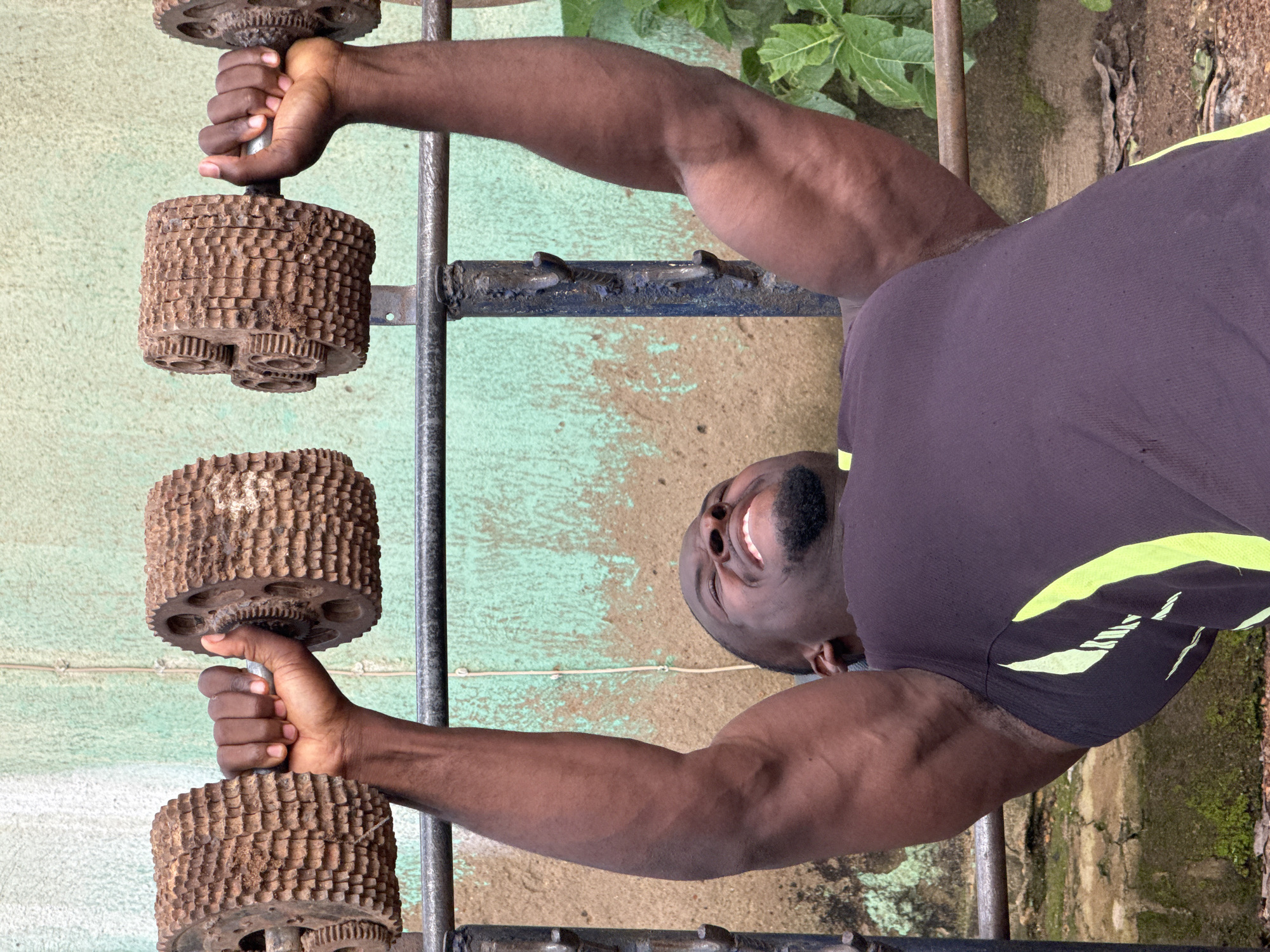 A muscular Black man with dark skin is performing a bench press exercise with two heavy, rustic dumbbells. He is lying on a bench, with his arms extended upwards, holding the dumbbells. His face is contorted with exertion, his eyes are closed, and his mouth is open. He is wearing a dark purple tank top with lime green accents on the side. The dumbbells are made of metal with a rough, textured surface resembling gears or cogs, and they appear to be antique or homemade. He is on a metal weight rack. The background consists of a wall with a faded, light green and brown textured paint, and some green foliage to the right. The setting appears to be an outdoor gym, possibly in a less developed area, with an unpaved ground surface and scattered debris. This scene is located in the city of Akwanga, Nigeria.
