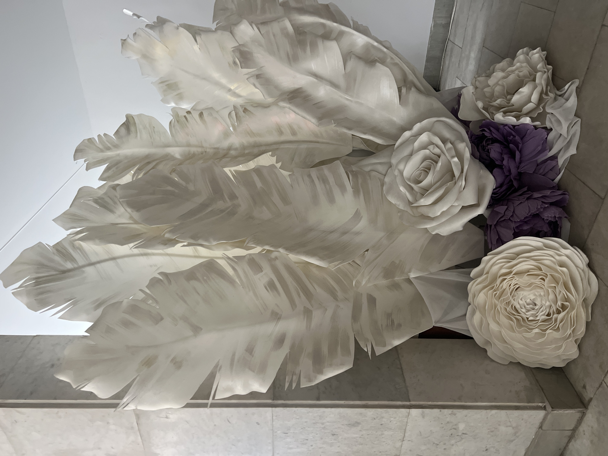 The image captures a static decorative arrangement composed of oversized artificial leaves and flowers, set against an indoor backdrop in Kyiv, Ukraine. The main subjects are large, fan-shaped leaves, predominantly white or off-white, filling the left and central portions of the frame. These leaves have a stylized, somewhat translucent appearance with visible brushstroke-like patterns, reminiscent of tropical foliage.
Interspersed among these leaves, primarily on the right side, are clusters of large artificial flowers. Prominent among them are white and off-white roses with layered petals, and several ruffled flowers in a striking dark purple or plum hue, possibly peonies or similar varieties. Another multi-petaled off-white flower, resembling a ranunculus or camellia, is visible towards the bottom right.
The background consists of a light-colored, possibly white or light grey, wall in the upper and left areas, transitioning to a light-toned tiled floor towards the bottom and right. The tiles appear to be square or rectangular with subtle variations, suggesting a marble or ceramic finish. The lighting is bright and even, characteristic of an interior space. The overall display conveys an elegant and decorative ambiance. No people or text are visible in the image.