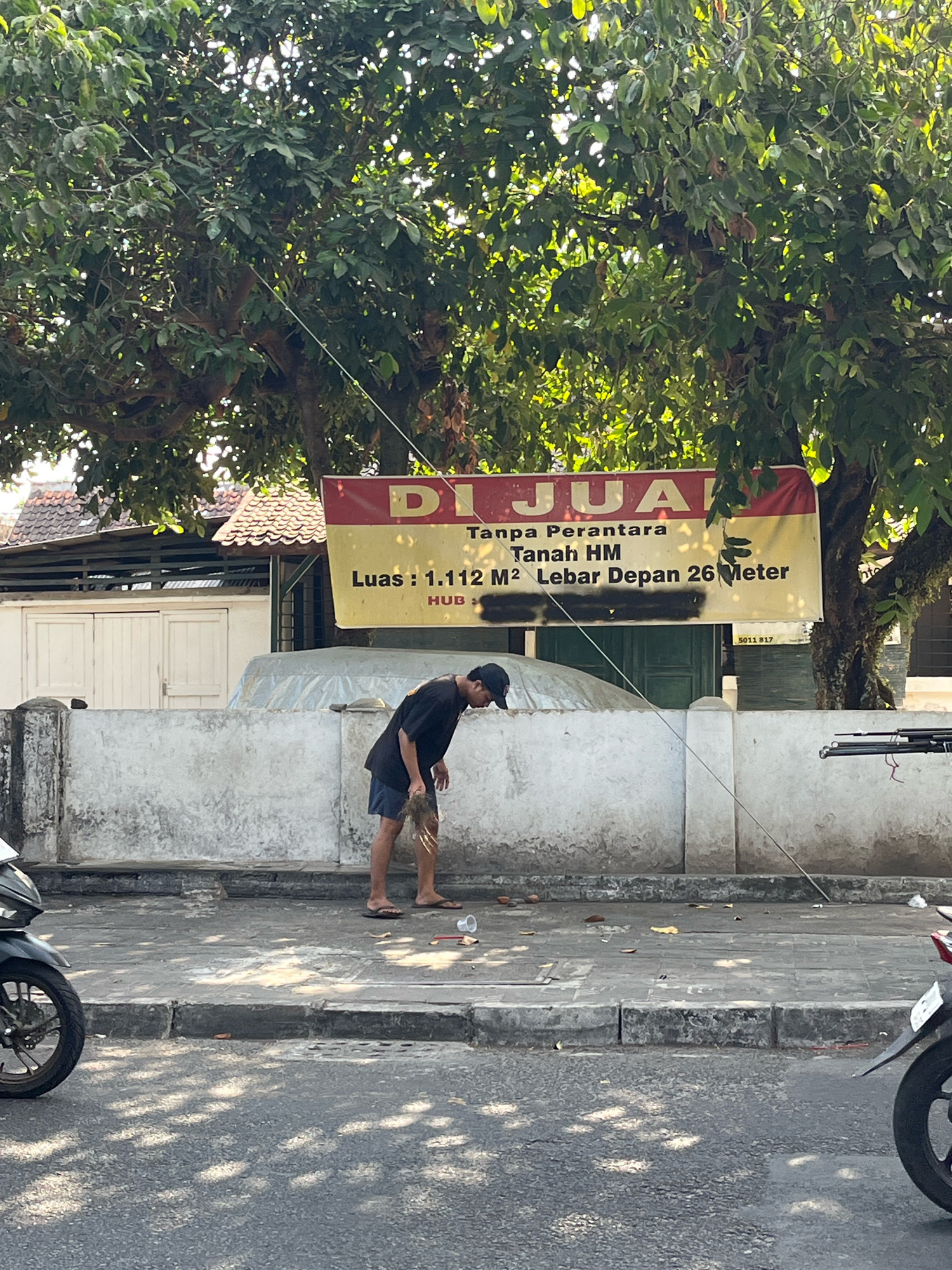A man wearing a black t-shirt and blue shorts bends down to pick up something from the ground on a sidewalk in front of a white wall. He is barefoot and carries a bunch of dried leaves in his right hand. A large yellow banner with red text is visible behind him, with the words "DIJUAL" (for sale) and property details. To his left, a motorcycle is parked. The sidewalk is paved with concrete blocks, and the street is visible in the foreground, with shadows from trees falling on it. A white wire extends from the upper left to the lower right of the frame, running diagonally across the scene. The environment appears to be a city street in Yogyakarta, Indonesia, with a house and trees in the background.
