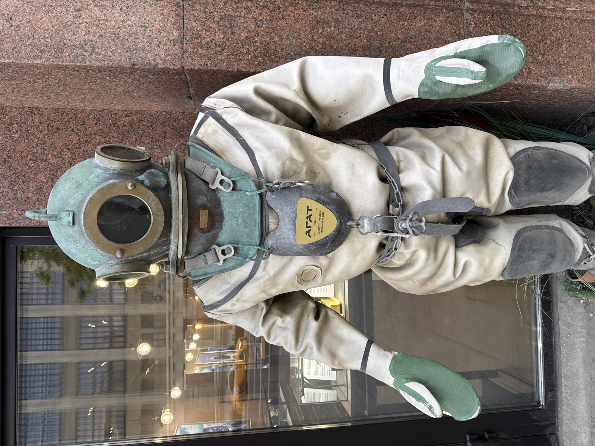 A full-bodied vintage diving suit with a copper helmet sits in front of a shop window. The suit is a pale, off-white color with green accents on the gloves and portions of the chest piece. The helmet is copper and shows signs of age and wear with a verdigris patina. A plate on the chest of the diving suit has text in Russian. The window behind the suit reflects a multi-story building. The photo appears to have been taken outdoors in front of a storefront. Based on the Russian text on the diving suit, the location is Moscow, Russia.