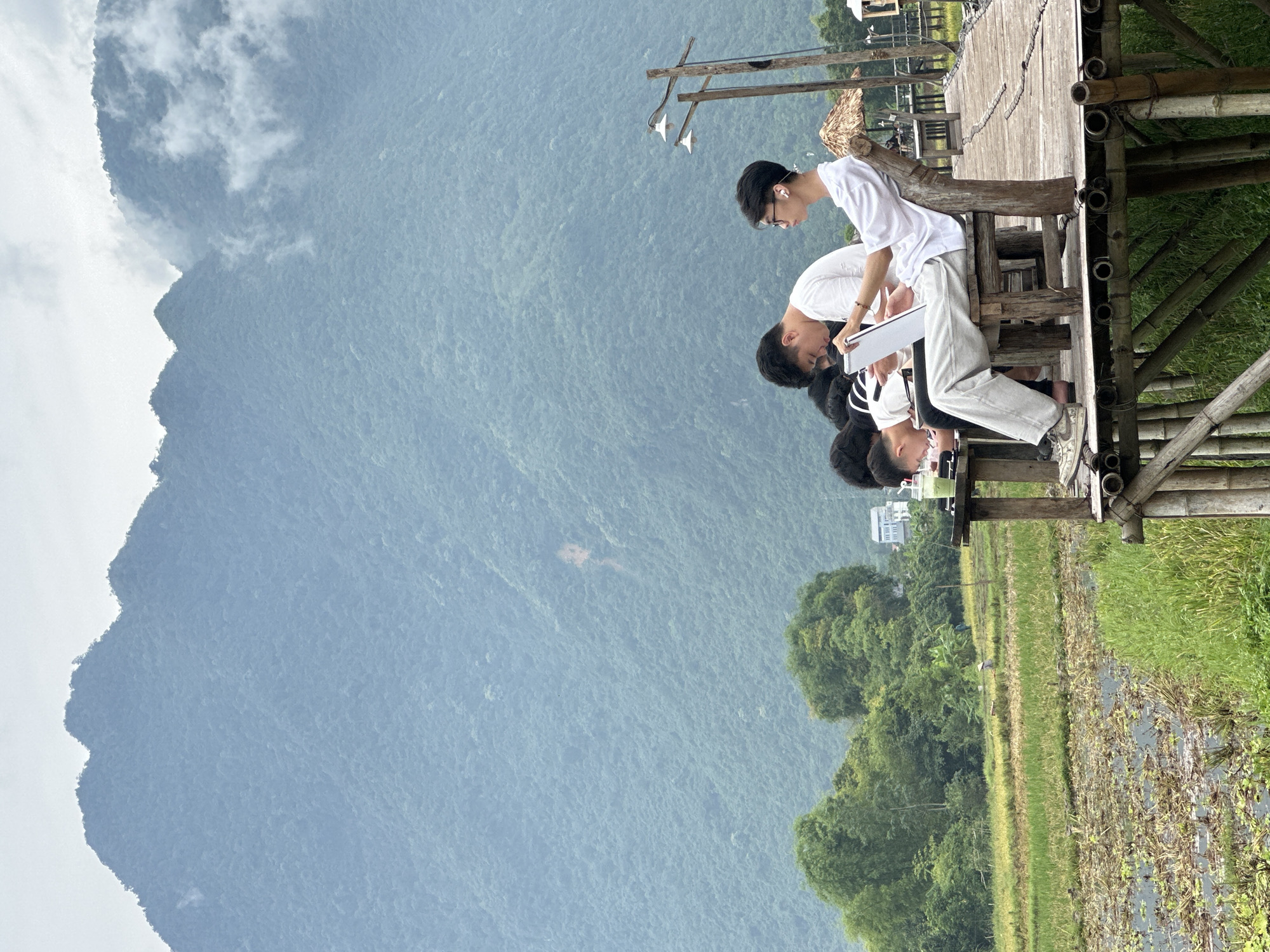 In Mai Chau, Vietnam, a group of young people sit on a wooden deck overlooking a lush green rice field with a mountainous backdrop. The individuals are engaged in various activities. One person is looking down at a phone, while another is focused on a tablet. A third person, wearing glasses and white earbuds, is holding a tablet. The deck itself is constructed from bamboo poles, with a rustic wooden railing and seating. Beyond the rice field, there are trees and a distant building. The sky is overcast, with some clouds obscuring the peaks of the imposing, tree-covered mountains.