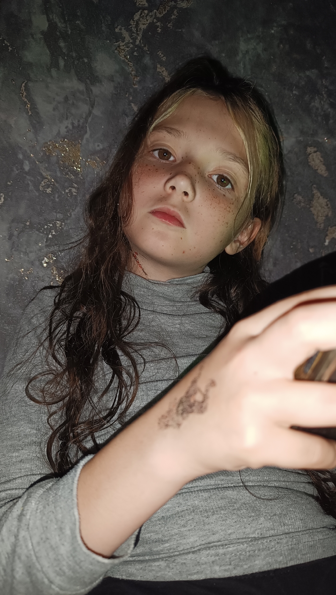 A close-up, chest-up portrait of a young person with light brown hair and freckles is shown. They are wearing a gray turtleneck shirt. The background is dark and textured, possibly a wall with an abstract pattern. The person's gaze is directed towards the camera. A tattoo or mark is visible on their left forearm. The lighting is dim, creating shadows.