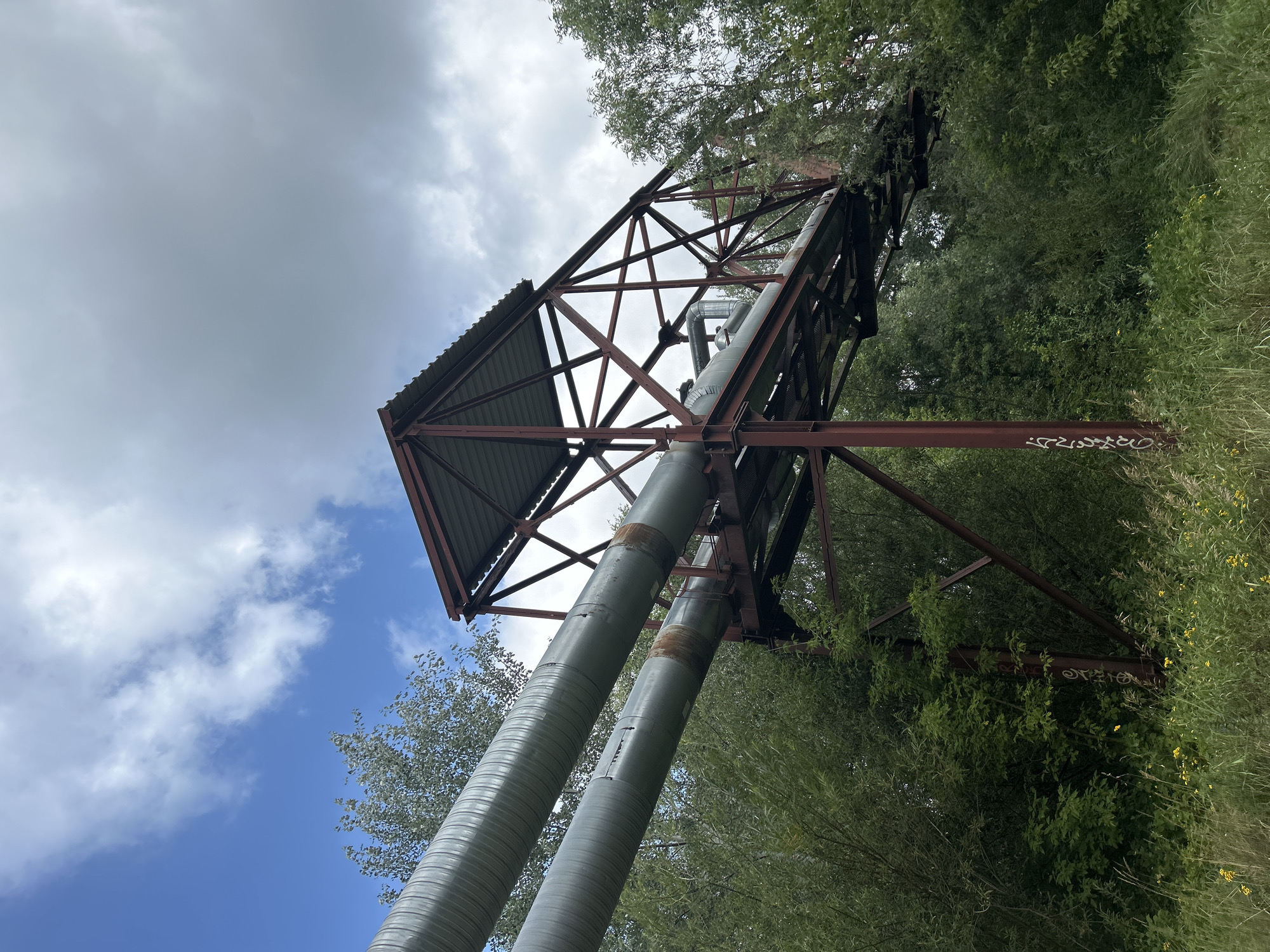 An industrial pipeline structure is visible in a natural environment. Two large, insulated pipes run horizontally across the frame, supported by a rusted metal framework. The framework has a corrugated metal roof over a section of the pipes. The surrounding area is overgrown with lush green trees, bushes, and tall grass. Small yellow flowers are scattered throughout the vegetation. The sky above is partly cloudy with a mix of blue sky and white clouds. The image appears to be taken from a low angle, looking up at the pipeline. There are no people visible in the scene.