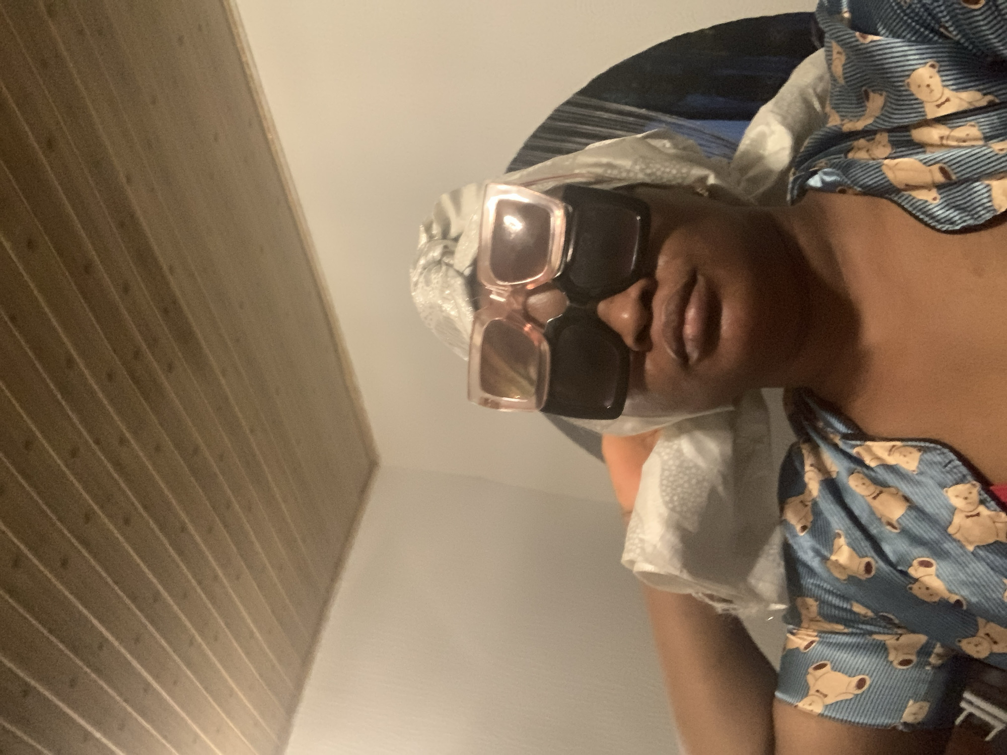 A medium shot of a woman wearing a white patterned head covering and large square sunglasses. She is wearing a blue shirt with a teddy bear print. The ceiling is visible and appears to be made of wood panels. The location is most likely inside a residence. There are no visible indicators that the location is Barakin Akawo, Nigeria.