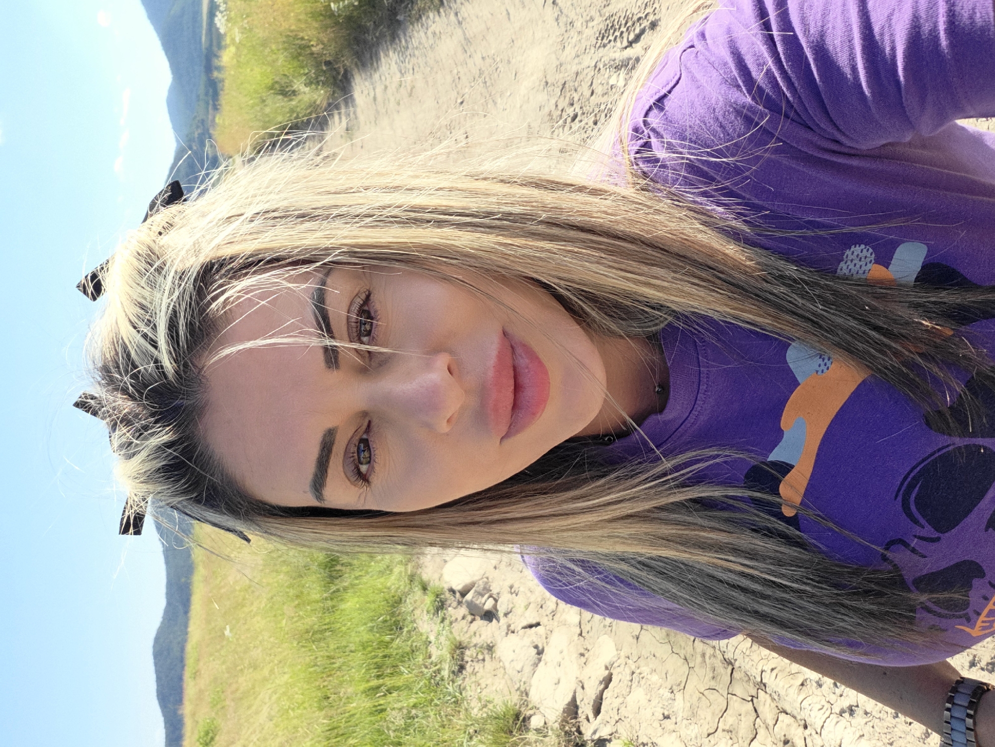 A woman with blonde hair, styled with hair clips at the top, is standing outdoors on a dirt path. She is wearing a purple t-shirt with a graphic design and a watch on her left wrist. The background features a field of dry grass, a dirt path, and distant hills under a clear blue sky. The scene appears to be located in the vicinity of Ravnogor, Bulgaria, suggested by the landscape.