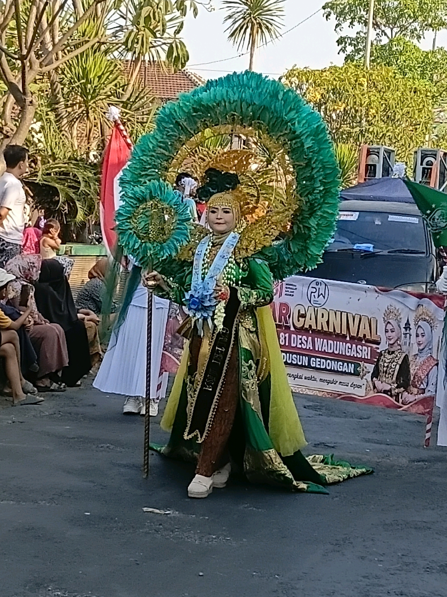 A person in a colorful, elaborate costume walks down a street in Wadungasri, Indonesia, during a carnival. The costume features a large, fan-like headpiece adorned with green and gold feathers, a green and gold dress with a long train, and a sash with blue flowers. The person holds a decorative staff in their right hand. In the background, a banner reads "KARNIVAL 81 DESA WADUNGASRI DUSUN GEDONGAN," indicating the location and event. Spectators are visible sitting on the side of the road, and a vehicle is parked behind the banner. The scene appears to be set during daytime, with sunlight illuminating the area.