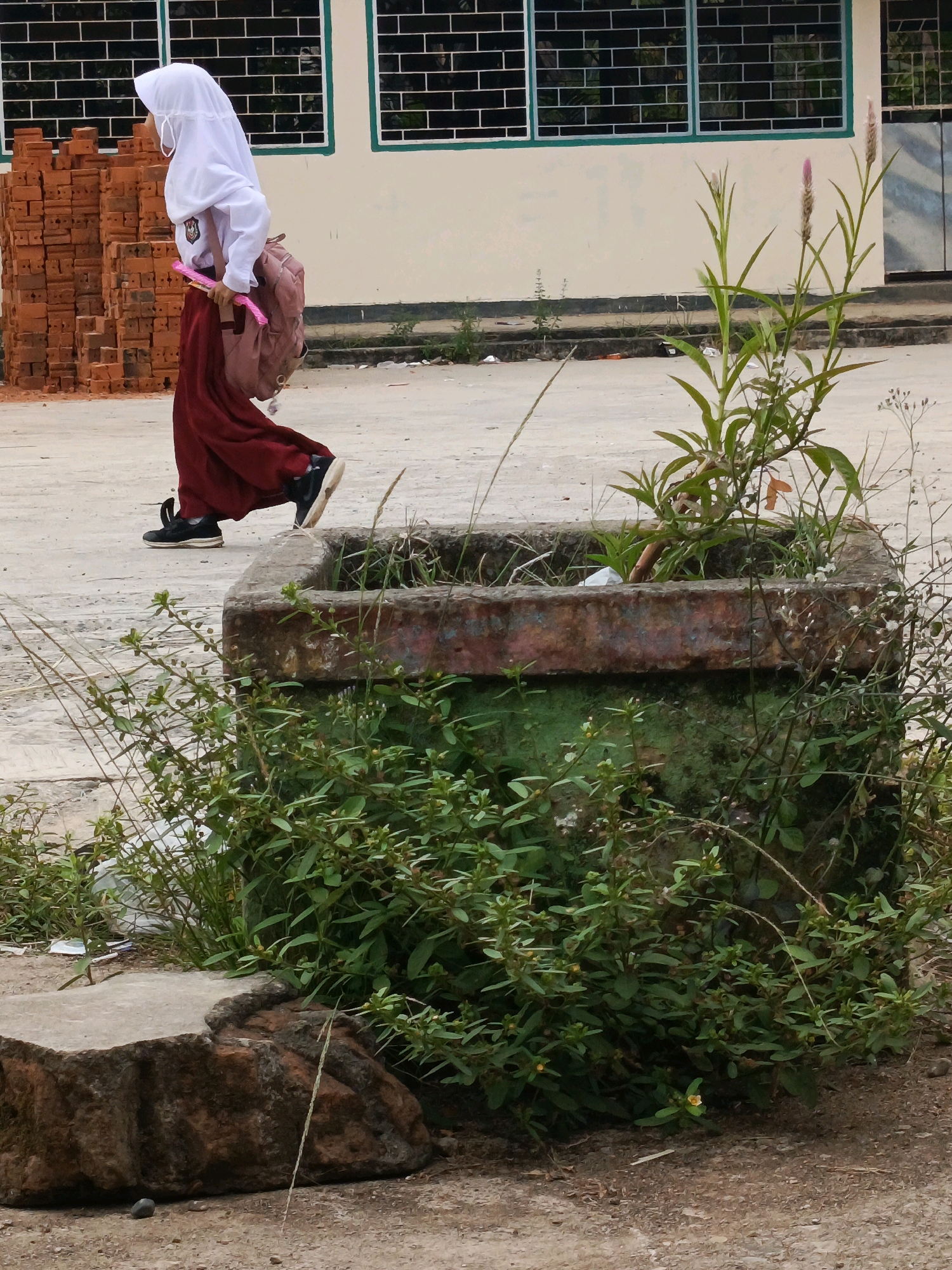A young person wearing a white hijab and school uniform, including a maroon skirt, walks outdoors past a stack of red bricks and a concrete planter filled with greenery. The individual carries a pink backpack and a pink object. The scene appears to be an outdoor area with a building featuring windows in the background. The ground is paved with concrete and dirt. This image was taken in Wonosari, Indonesia.