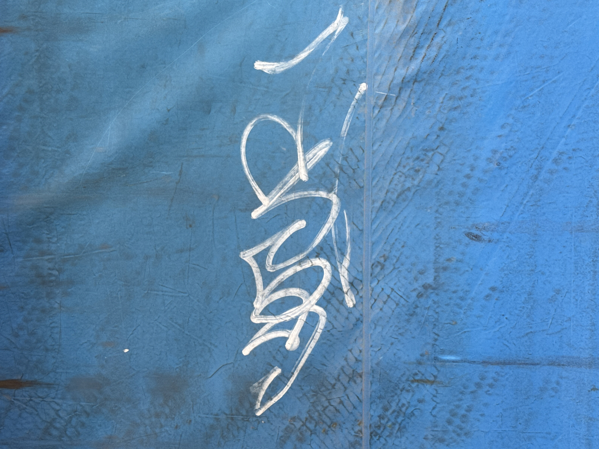 This media file features a close-up, eye-level shot of a blue, textured surface with white graffiti. The graffiti appears to be a tag, written vertically in a stylized, script-like font. The characters are fluid and abstract, making them difficult to definitively decipher. The surface itself has a weathered appearance, with visible scratches, scuffs, and an uneven texture that suggests it might be an outdoor surface like a painted wall or a metal sheeting. The lighting appears to be natural, casting subtle shadows and highlighting the texture of the surface. There are no people, specific objects, or clear scene elements visible beyond the graffiti and the blue background. The context of Karon, Thailand, does not provide specific visual cues within the image itself.