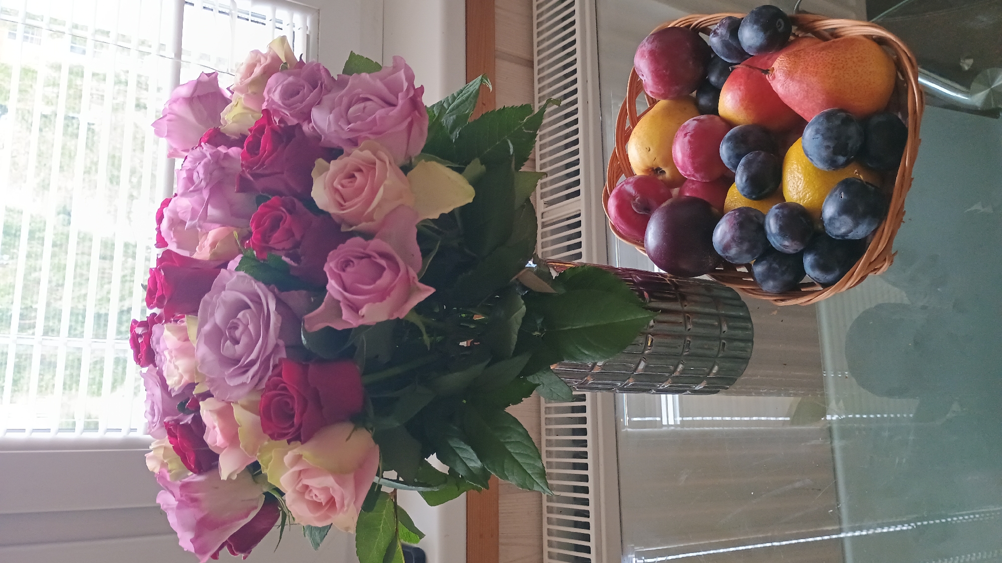 A bouquet of roses in various shades of pink, red, and lavender is displayed in a metallic vase on a glass table. To the right of the vase, a woven basket contains a selection of fruits, including plums, lemons, and pears. The scene is set against a backdrop of a window with blinds, and a radiator can be seen beneath the window. This image was taken in Sokolov, Czechia.