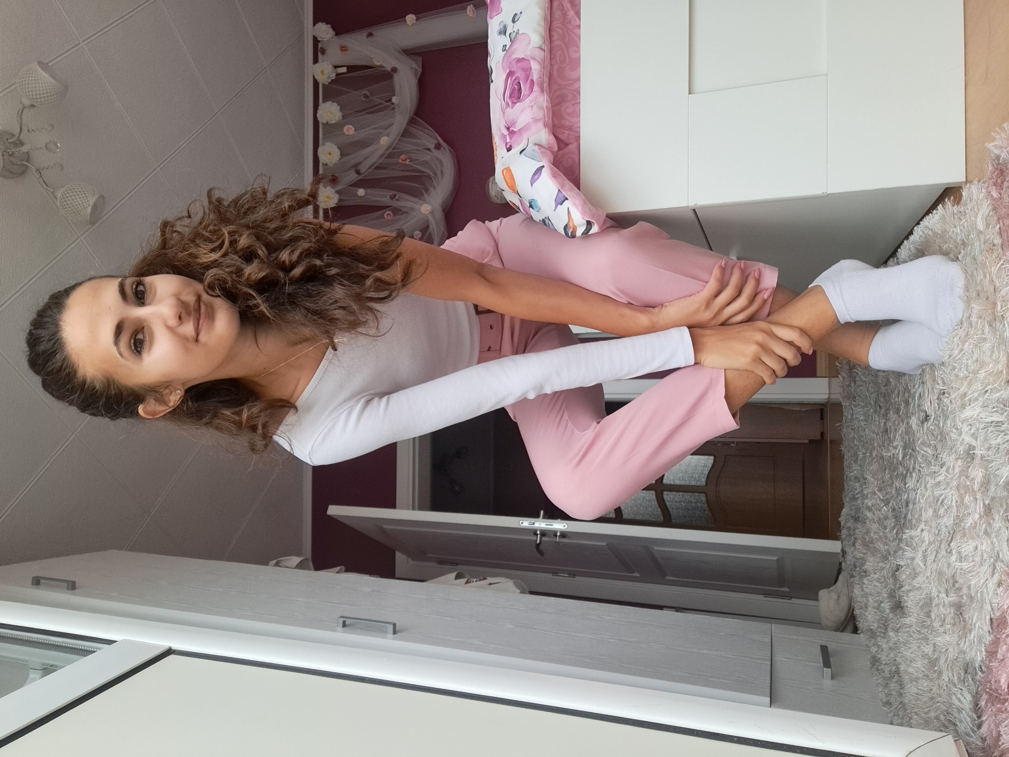 A young woman with curly brown hair pulled back in a ponytail is sitting on the edge of a white bed in a bedroom. She is wearing a white long-sleeved shirt, pink pants, and white socks. Her hands are clasped around her knees. The bed has a pink and white floral duvet. Behind the bed, there is a wall with a dark pink color, decorated with a sheer white canopy with small flowers. To the left of the bed is a white cabinet. A large fluffy white rug covers the floor in front of the bed and extends towards a doorway. The doorway leads to a hallway with a wooden door. The room appears to be well-lit by natural light from an unseen window.