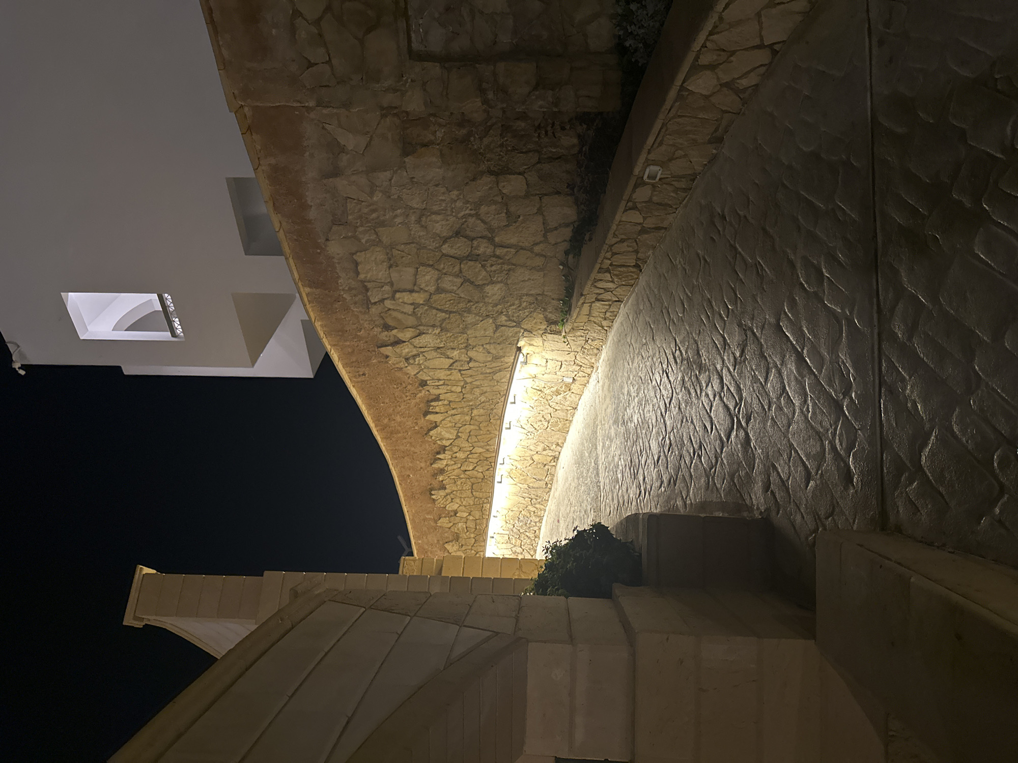 The image displays an outdoor, paved pathway at night, illuminated by artificial light. The pathway, constructed from dark, textured paving stones patterned to resemble cobblestones, curves to the right and recedes into the distance.
To the right of the path, a prominent, high, and curved retaining wall extends along the curvature. This wall is constructed from irregularly shaped, natural-toned stones with hues of beige, brown, and orange. A continuous linear light fixture, emitting a warm, yellow-toned light, is mounted horizontally along the base of this stone wall, illuminating the pathway and the lower section of the wall. Above this light strip, a narrow planter containing sparse, dark vegetation runs along the wall. Further along the wall on the right, a dark, rectangular recess or opening is visible.
Above the stone retaining wall, against a uniformly dark night sky, a modern, multi-story building with clean, white facades is visible. A distinctive rectangular window opening on the upper part of this building reveals an interior space with an arched ceiling and a horizontal balustrade. Other darker, rectangular recesses are present on the lower visible sections of the white building.
To the left of the path, a substantial architectural structure, built from light-colored, coursed masonry blocks, forms a partial archway or pillar in the foreground. A dark green shrub is situated at the base of this structure, adjacent to the path. No individuals are present in the scene.
Location: Unknown, Unknown.