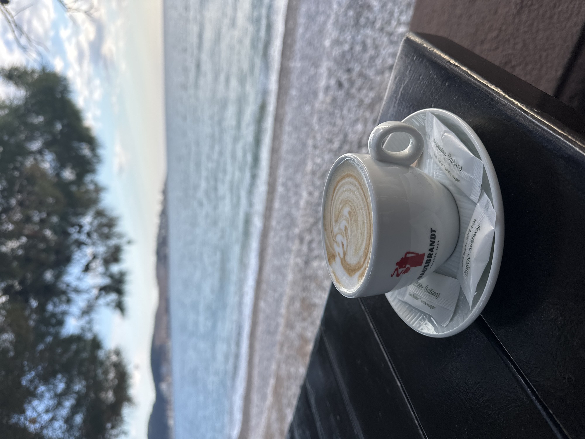 A cup of Hausbrandt latte sits on a saucer with sugar packets labeled "Šušanj" on a dark, possibly wooden, table. The background is a coastal view with the sea, beach, trees, and cloudy sky suggesting the location is Šušanj, Montenegro.