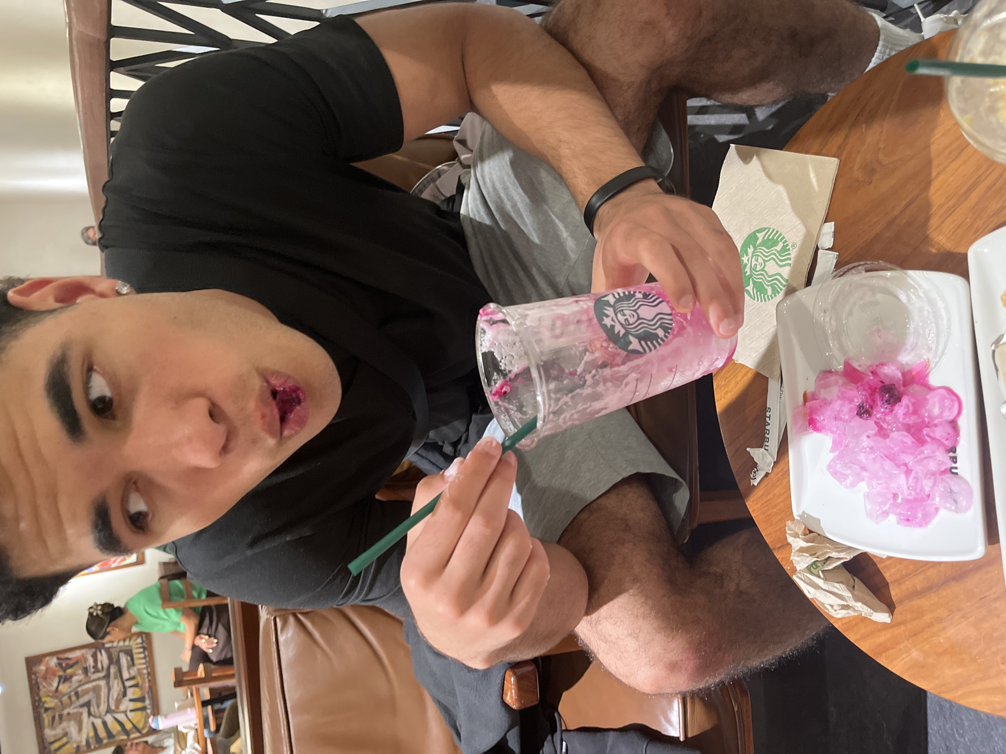 A person is seen close-up, holding a Starbucks cup with pink residue and a green straw. The person has pink residue around their mouth, suggesting they have been consuming the drink. Next to the cup on a wooden table is a Starbucks napkin and a white plate with remnants of a pink substance. Other people can be seen in the background. The setting is a Starbucks store, possibly in Puebla, Mexico.