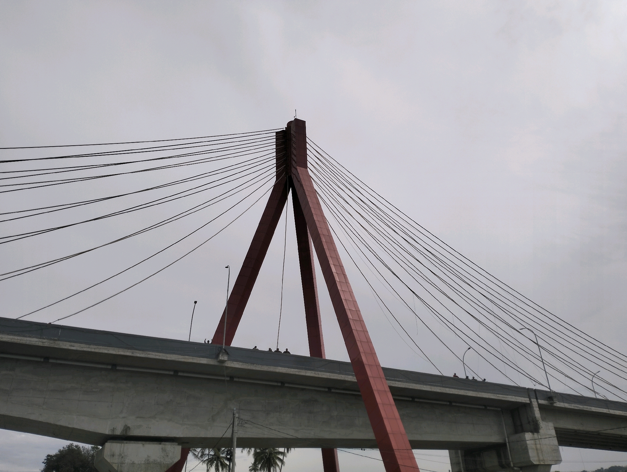 A view from below shows the underside of a concrete bridge supported by massive red pylons and a network of suspension cables. The sky is overcast and gray. Several people, appearing as small silhouettes, are visible standing on the bridge deck near the pylons. There are also palm trees visible in the lower part of the image, suggesting a tropical or subtropical location. The scene is of the Siogung-Ogung bridge in Siogung-Ogung, Indonesia.
