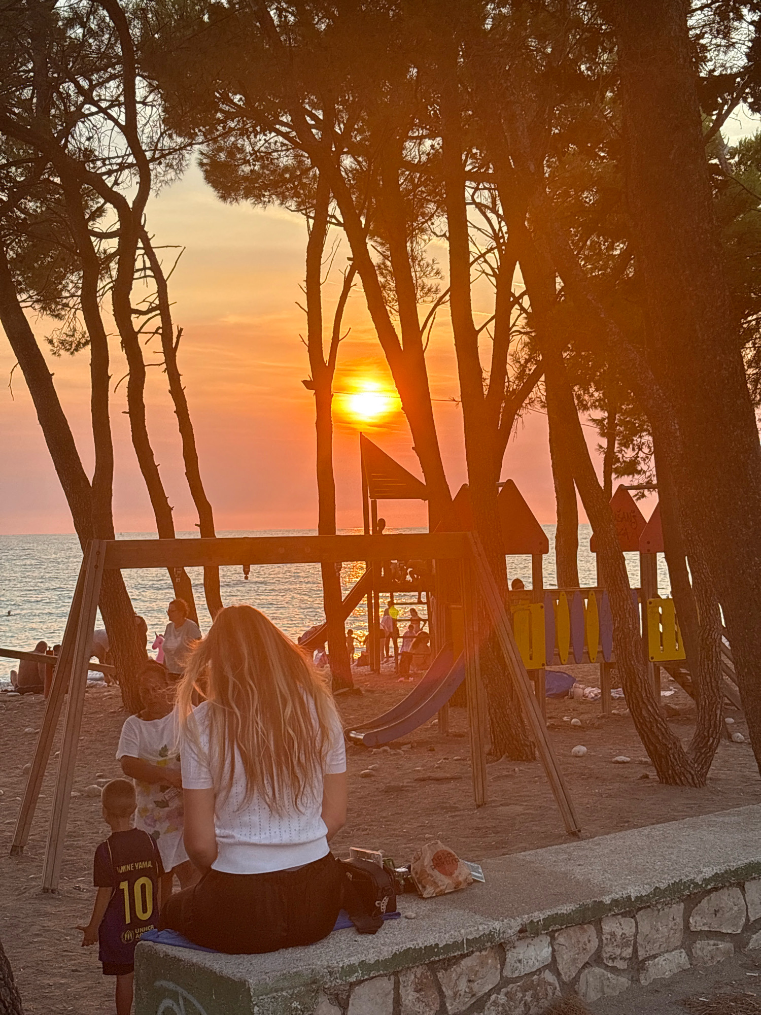 The image depicts a beach scene during sunset, likely in the city of Bar, Montenegro, given the presence of pine trees commonly found along the Adriatic coast and the distinctive playgrounds. The sky is a vibrant orange and yellow, with the sun low on the horizon, casting a warm glow over the scene.
In the foreground, a woman with long blonde hair is seated on a concrete wall, facing away from the camera. To her left, a young boy wearing a dark t-shirt with the number "10" and the UNHCR logo stands. Behind them, partially obscured by trees and a swing set, a playground with a slide and colorful panels is visible.
Several other individuals are scattered throughout the scene, some near the playground and others on the beach closer to the water. The beach itself appears to be sandy, with scattered rocks and vegetation. The sea is calm, reflecting the colors of the sunset. The overall atmosphere is relaxed and peaceful, suggesting a family-friendly environment.