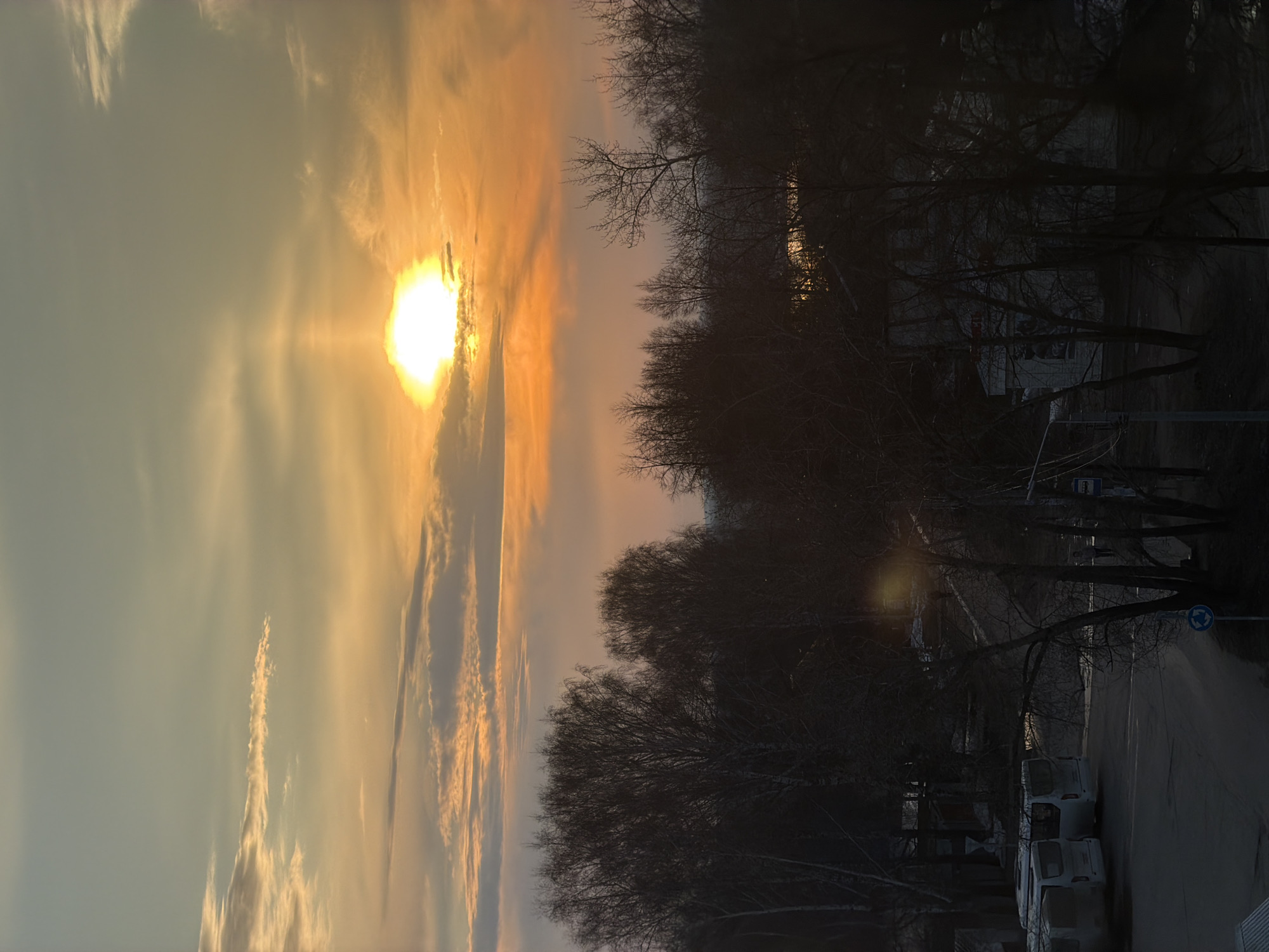 The image captures a scene in the city of Chaikovsky, Russia, during a sunset. The sun is positioned on the left, partially obscured by clouds, casting an orange and yellow glow across the sky. The right side of the image is dominated by silhouetted trees, indicating the presence of a park or wooded area along a street. Below the trees is a row of parked delivery trucks. A street light pole is visible near the center, and a roundabout sign is present further down the street. The overall scene suggests a quiet evening in a residential or commercial area of the city.