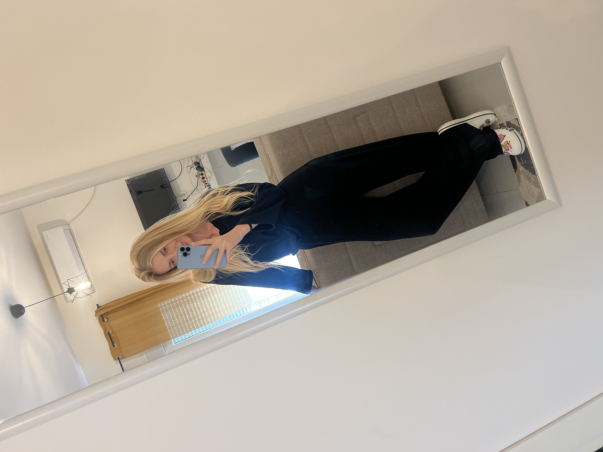 An upright mirror selfie captures a person with long blonde hair, dressed in a black long-sleeved top and black wide-leg pants, holding a light blue smartphone to take the photo. The person is wearing white platform shoes with colorful, cartoonish graphics, including a pink heart-shaped character and partially visible text "CRCC" and "ocs," suggesting they are Crocs brand footwear.
The reflection in the white-framed, full-length mirror reveals a contemporary indoor setting. Visible elements include a light grey fabric sofa or bed, a wall-mounted air conditioner unit, a television or monitor with associated cables, and a window fitted with a light brown roller blind or shutter. A modern pendant light fixture with a geometric cage design hangs from the ceiling. The room features light-colored walls, indicative of a bright, daytime setting. The photo was taken in Budva, Montenegro.