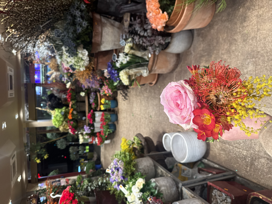 A flower shop interior displays an array of potted plants and cut flowers. A pink rose bouquet, featuring red and yellow accents, dominates the foreground. Ceramic vases and planters are scattered across the scene. The background showcases rows of colorful flowers, creating a vibrant and immersive shopping experience.